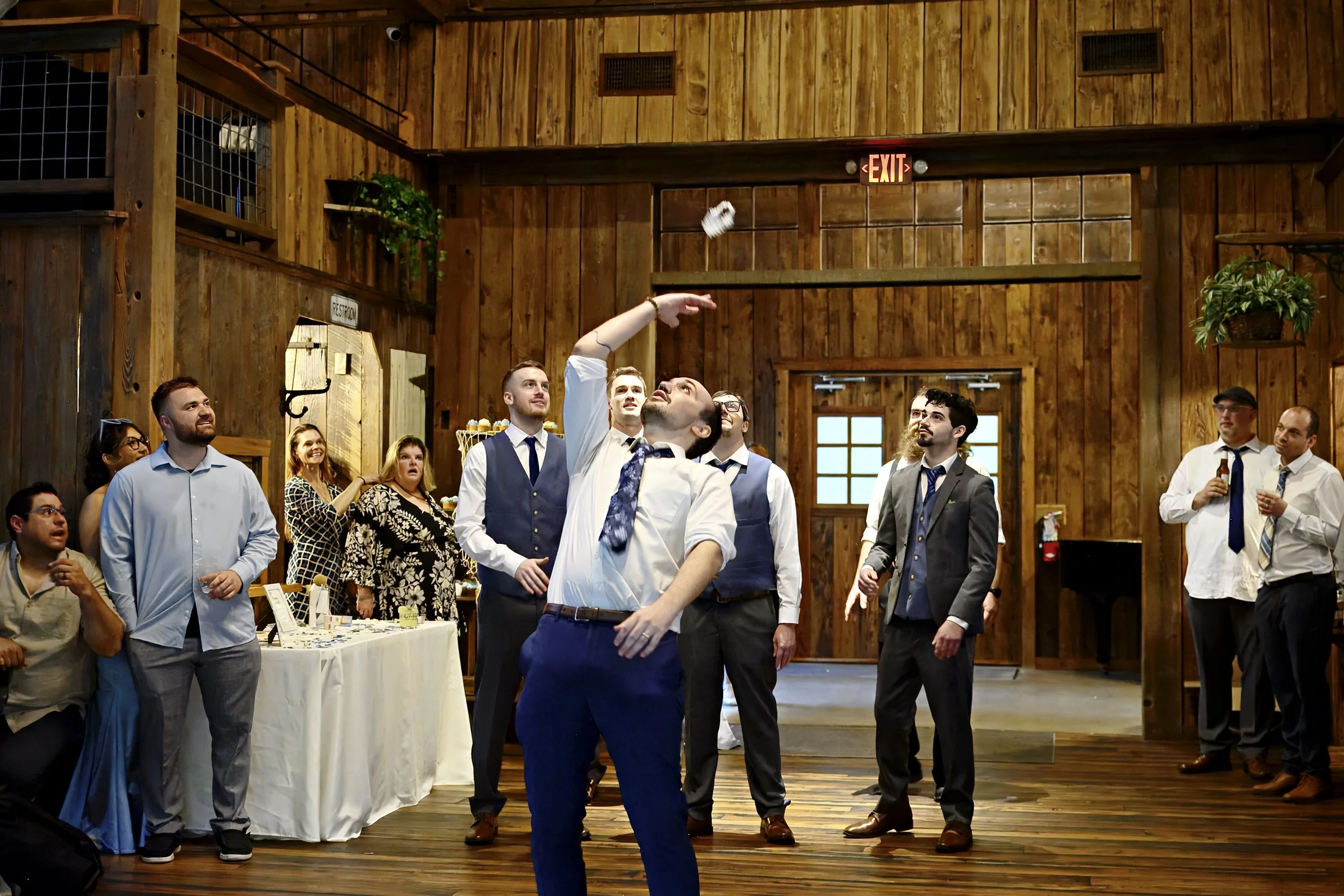 Group of people dressed in formal attire, some in suits and others in dresses, standing inside a rustic wooden building during a social event. One person in the center is tossing a small object into the air. Some people are observing and smiling. The