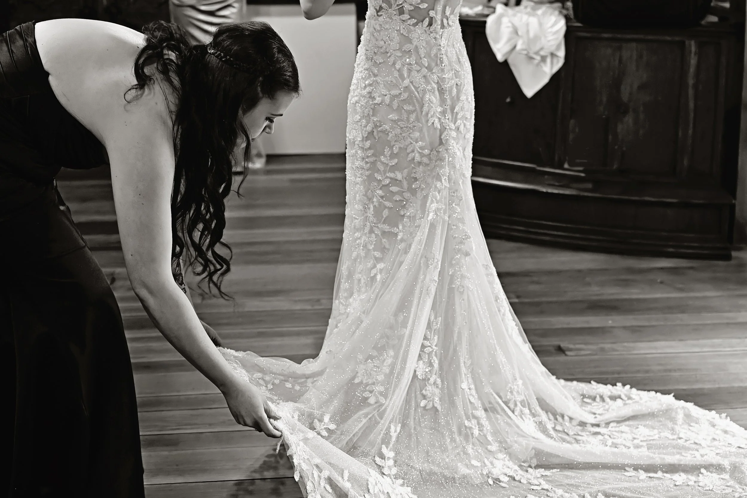 A woman in a black dress is bent down, holding the train of a wedding dress that another person is wearing. The wedding dress is detailed with lace and beads, and the scene takes place indoors on a wooden floor with a dark wooden cabinet in the backg