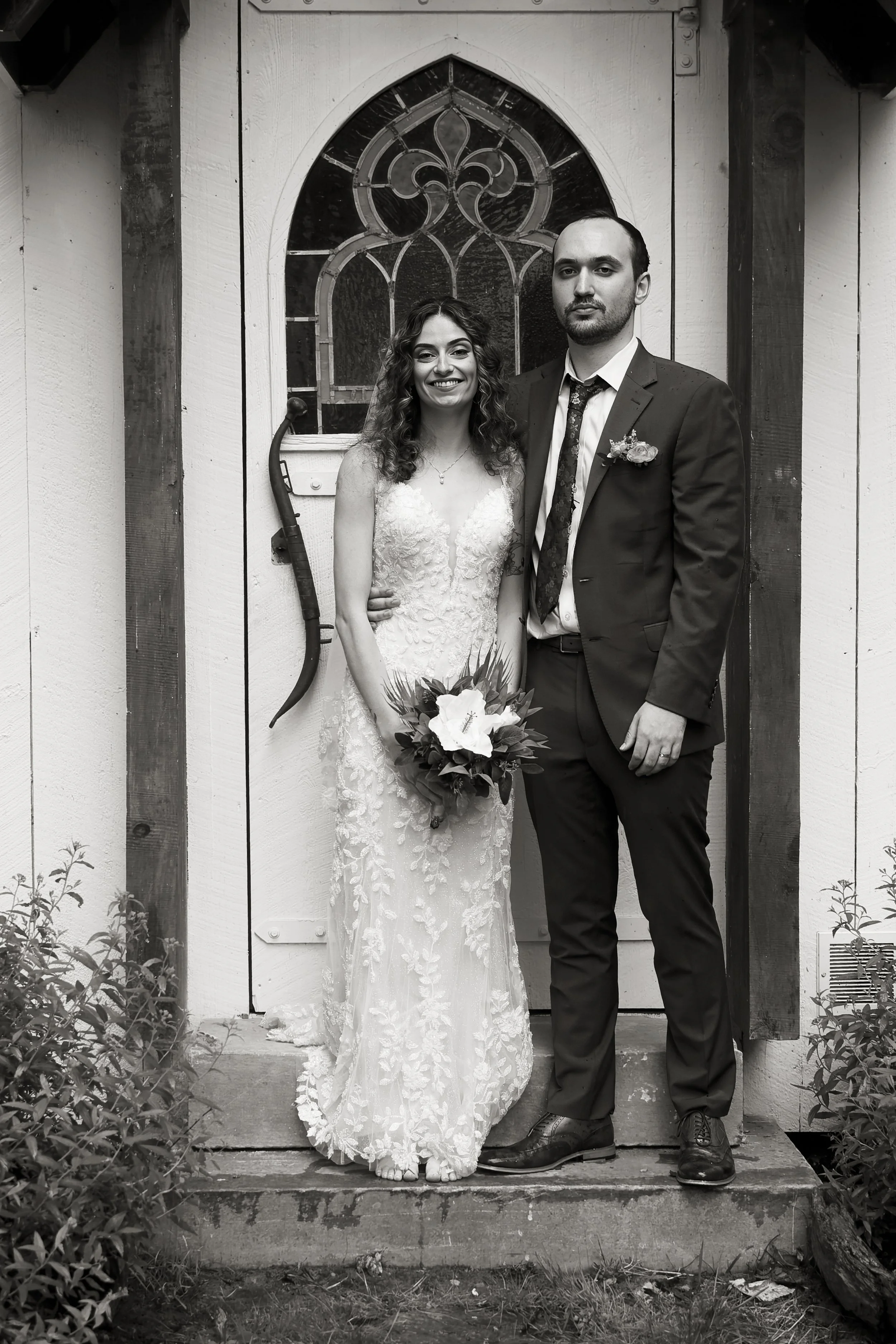 Black and white photo of a bride and groom standing in front of a small wooden building with a gothic style window. The bride is on the left, smiling, wearing a lace wedding dress and holding a bouquet of flowers. The groom is on the right, with a se
