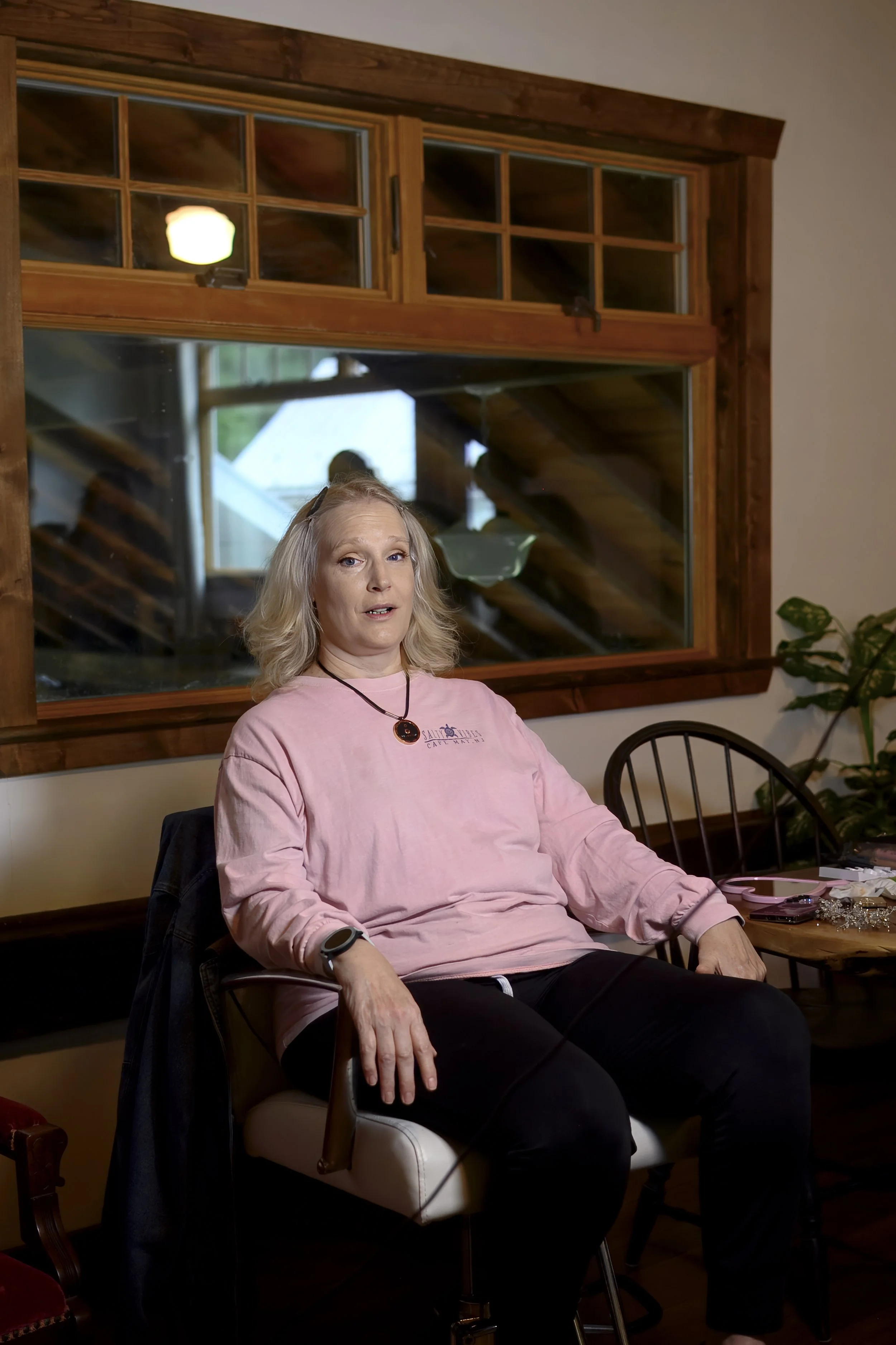 A woman with shoulder-length blonde hair, wearing a pink long sleeve shirt, sitting in a room with wooden framed windows, a round wooden table, and a plant in the corner.