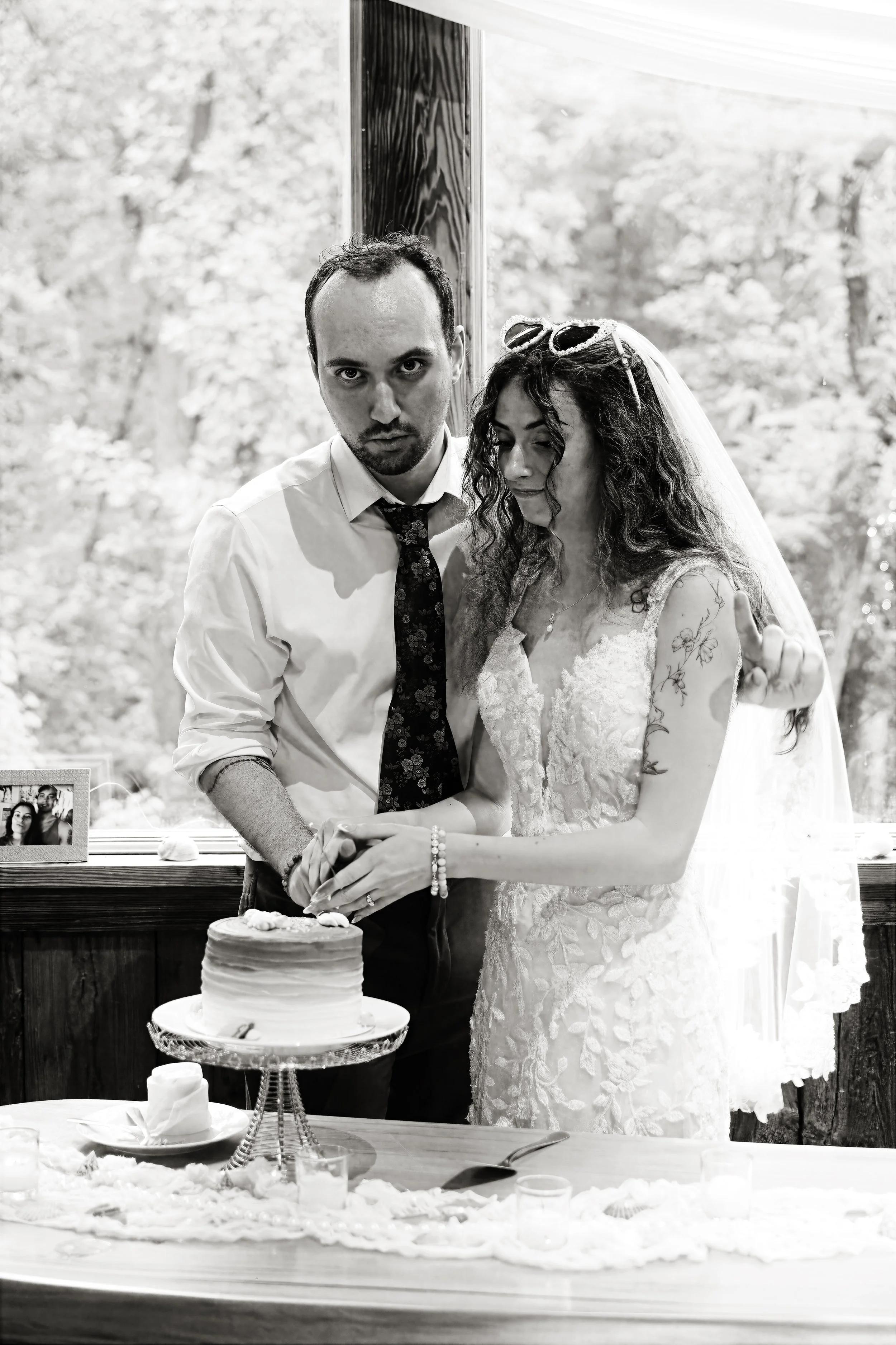 A bride and groom cutting a wedding cake together at their wedding reception, with a framed photo on the table and a wooded outdoor scene visible through a window behind them.