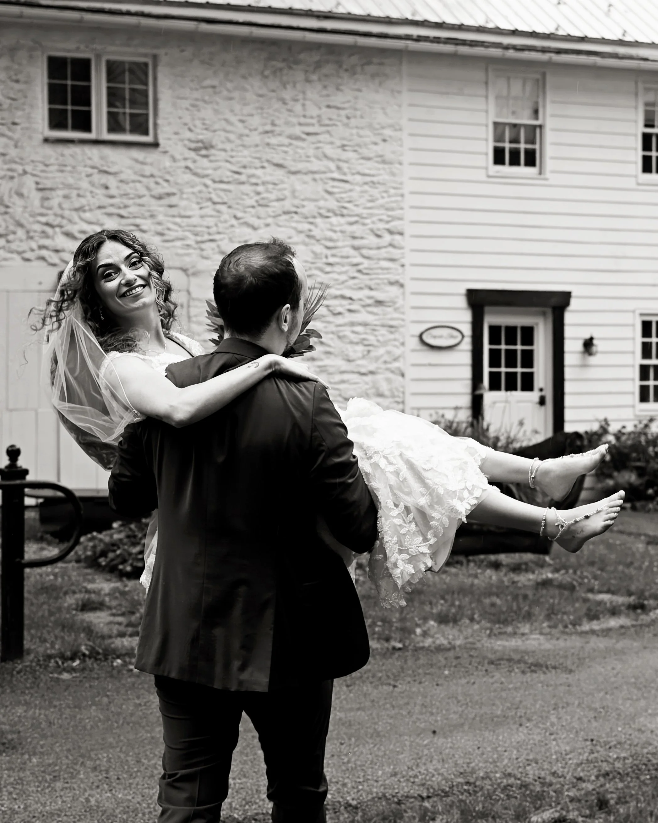 A woman in a wedding dress being carried by a man outdoors in front of a house, smiling and looking at the camera.