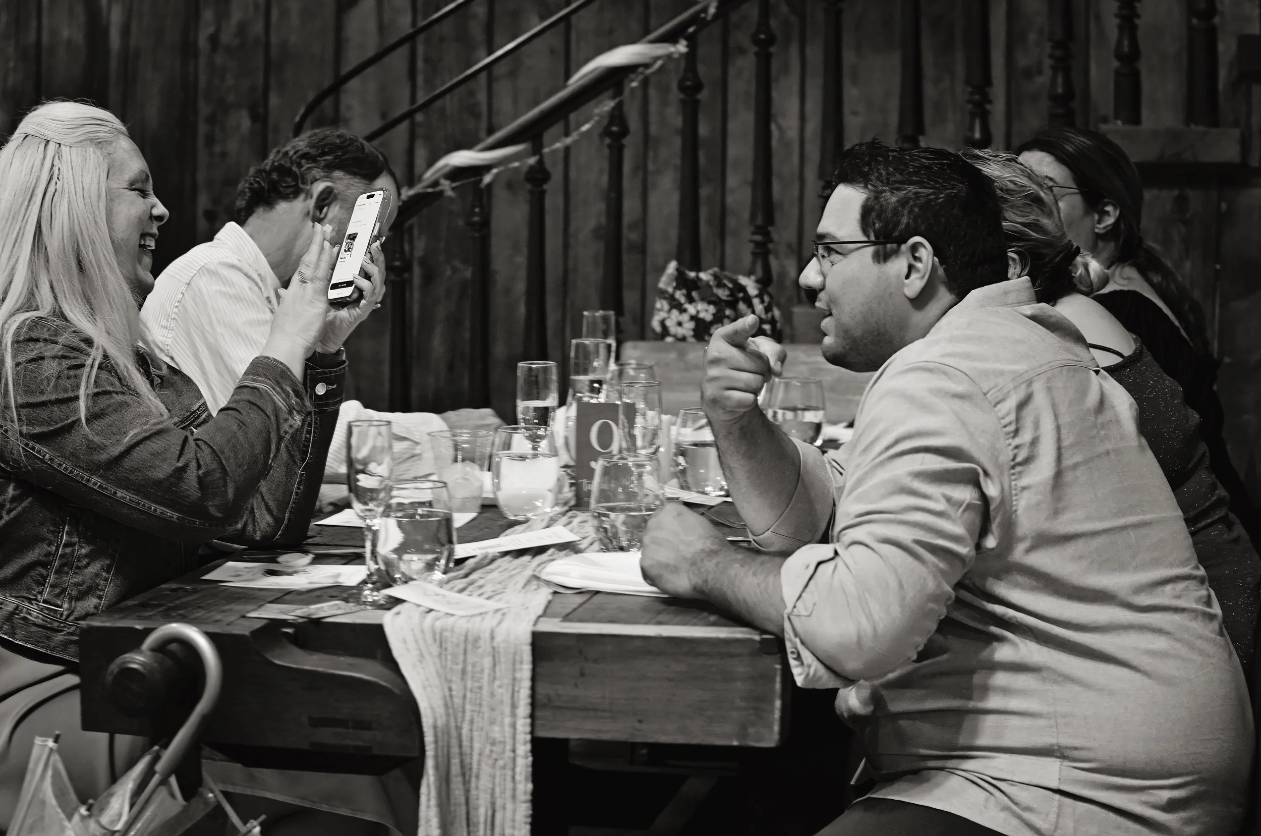A group of five people sitting at a dining table engaged in conversation, with glasses and utensils on the table, in a rustic setting with wooden walls and staircase in the background.