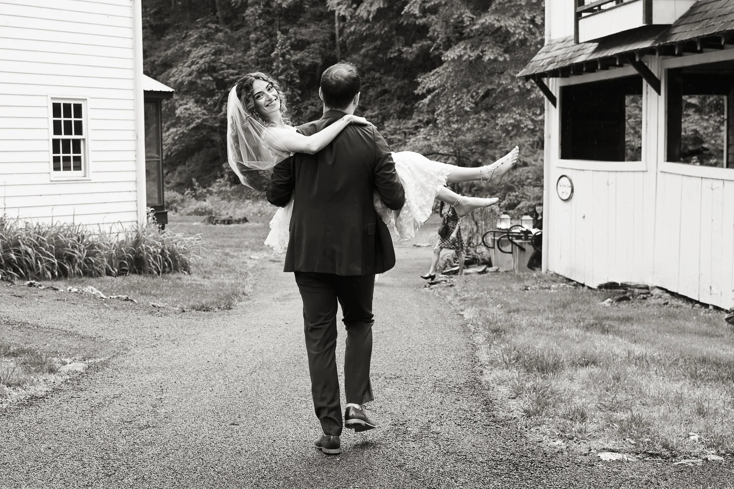 A groom in a suit is carrying a smiling bride in a wedding dress and veil, walking outdoors on a paved path with grass and flowers on either side. In the background, there are trees, a white house with siding, and a small wooden structure.