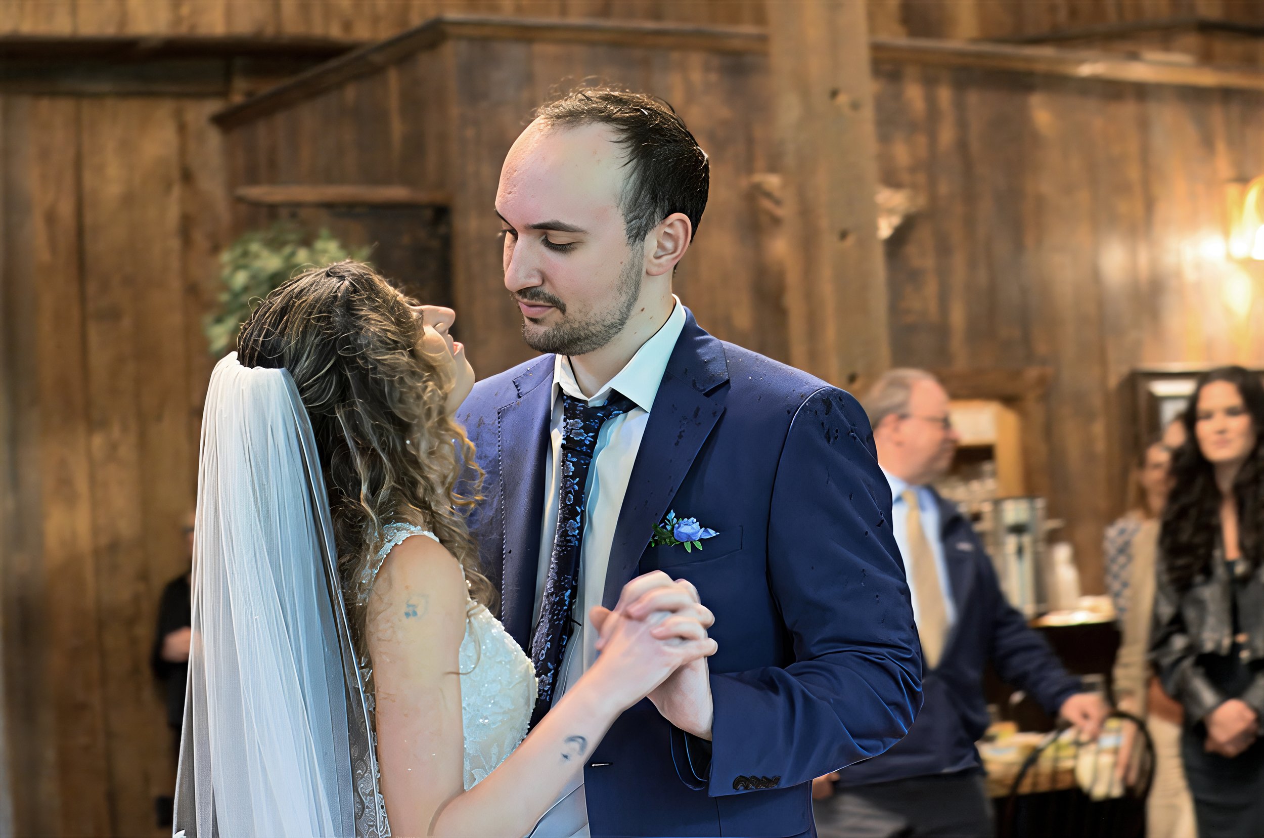 A bride and groom dance at their wedding reception in a rustic wooden venue with guests watching in the background.