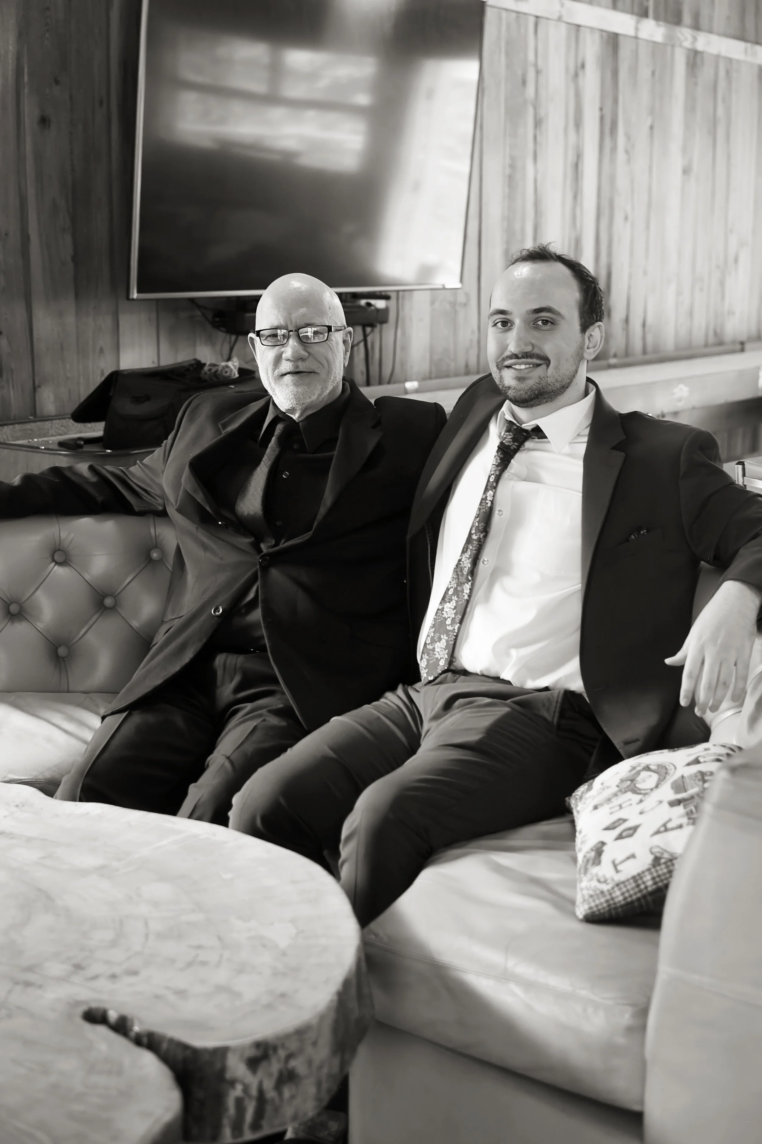 Two men in suits sitting on a tufted leather couch in a room with wood-paneled walls. A large flat-screen TV is mounted on the wall behind them. One man is bald and wearing glasses, the other has short dark hair. Both are smiling and looking at the c