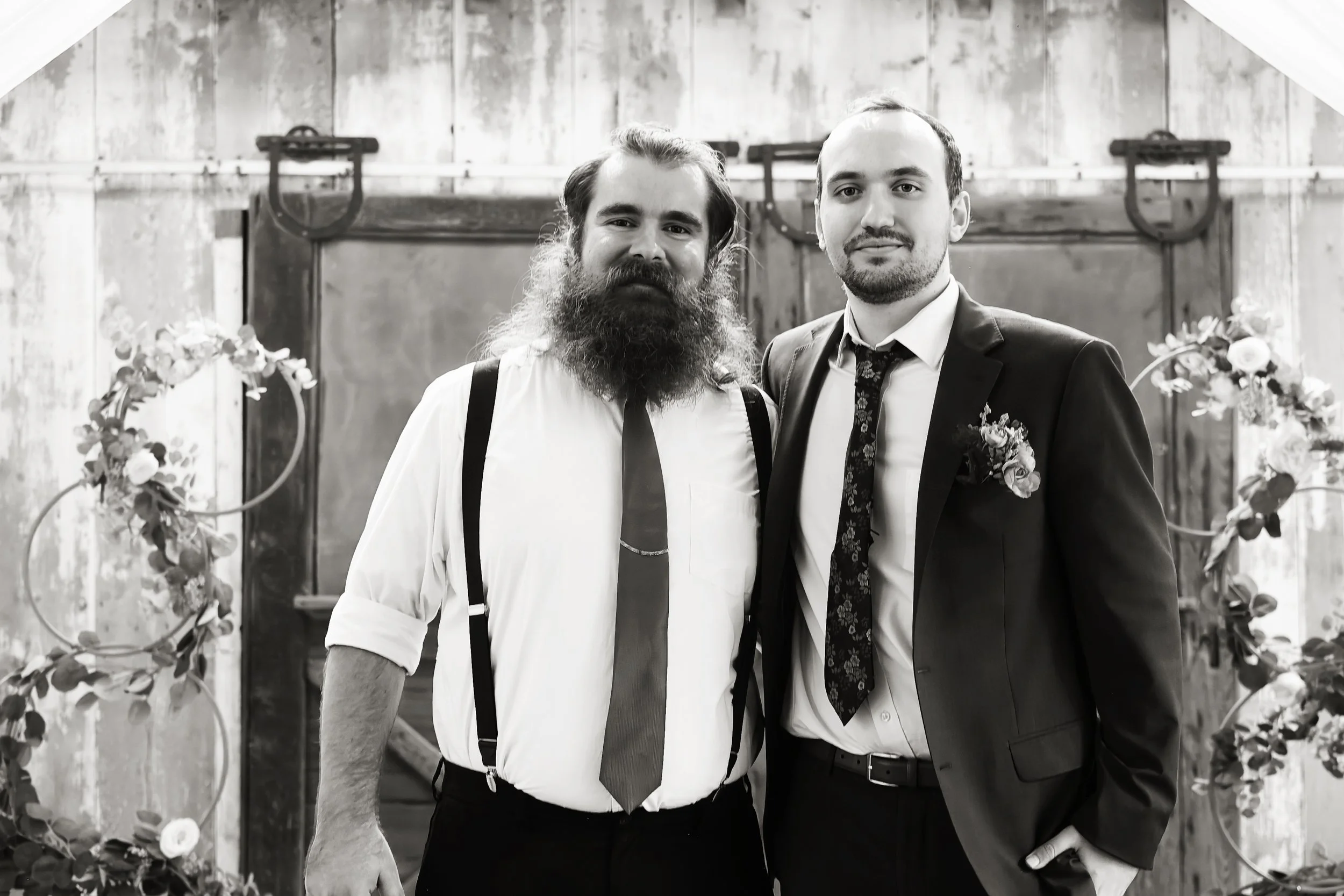 Two men dressed in formal attire standing close together, smiling at the camera. The man on the left has a full beard and is wearing suspenders, while the man on the right is clean-shaven and wearing a suit with a floral tie and boutonniere. They are