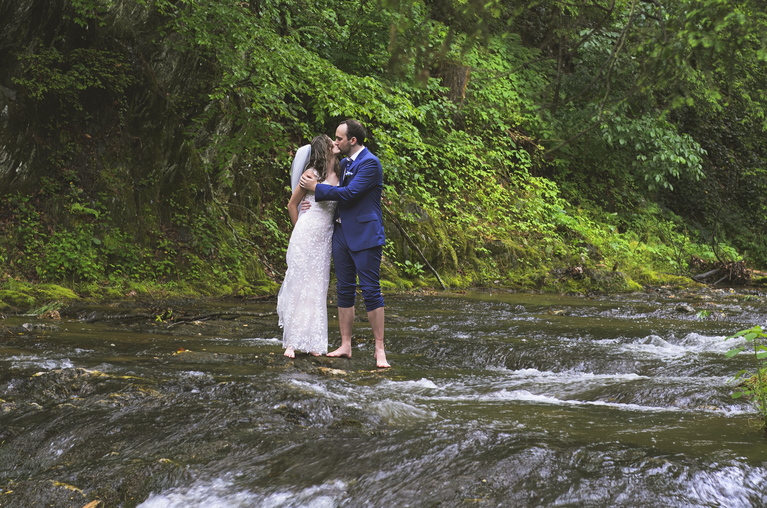 A bride and groom share a kiss while standing barefoot in a shallow stream surrounded by lush green foliage.