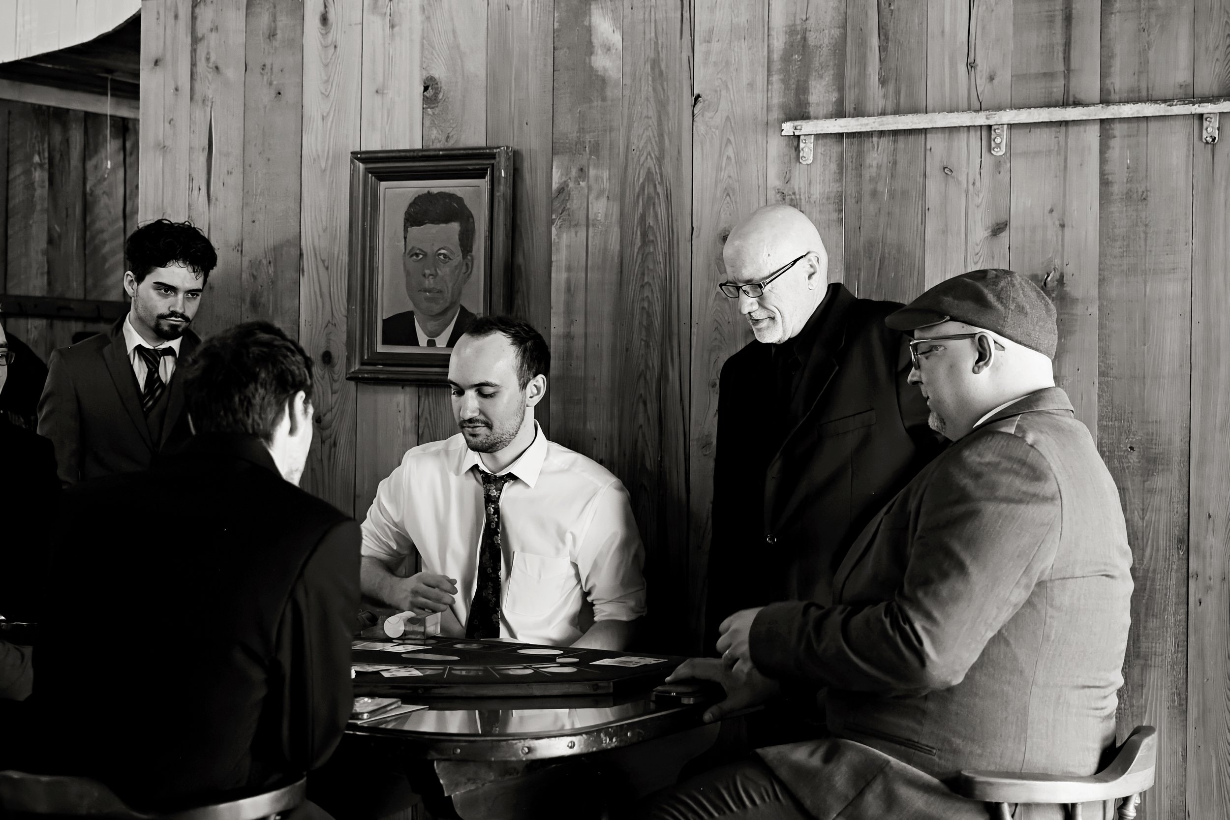 A group of five men gathered around a table playing a card game in a wooden room. One player is wearing a white shirt and a patterned tie, while others are in dark suits and glasses. There is a portrait of a man on the wall behind them.