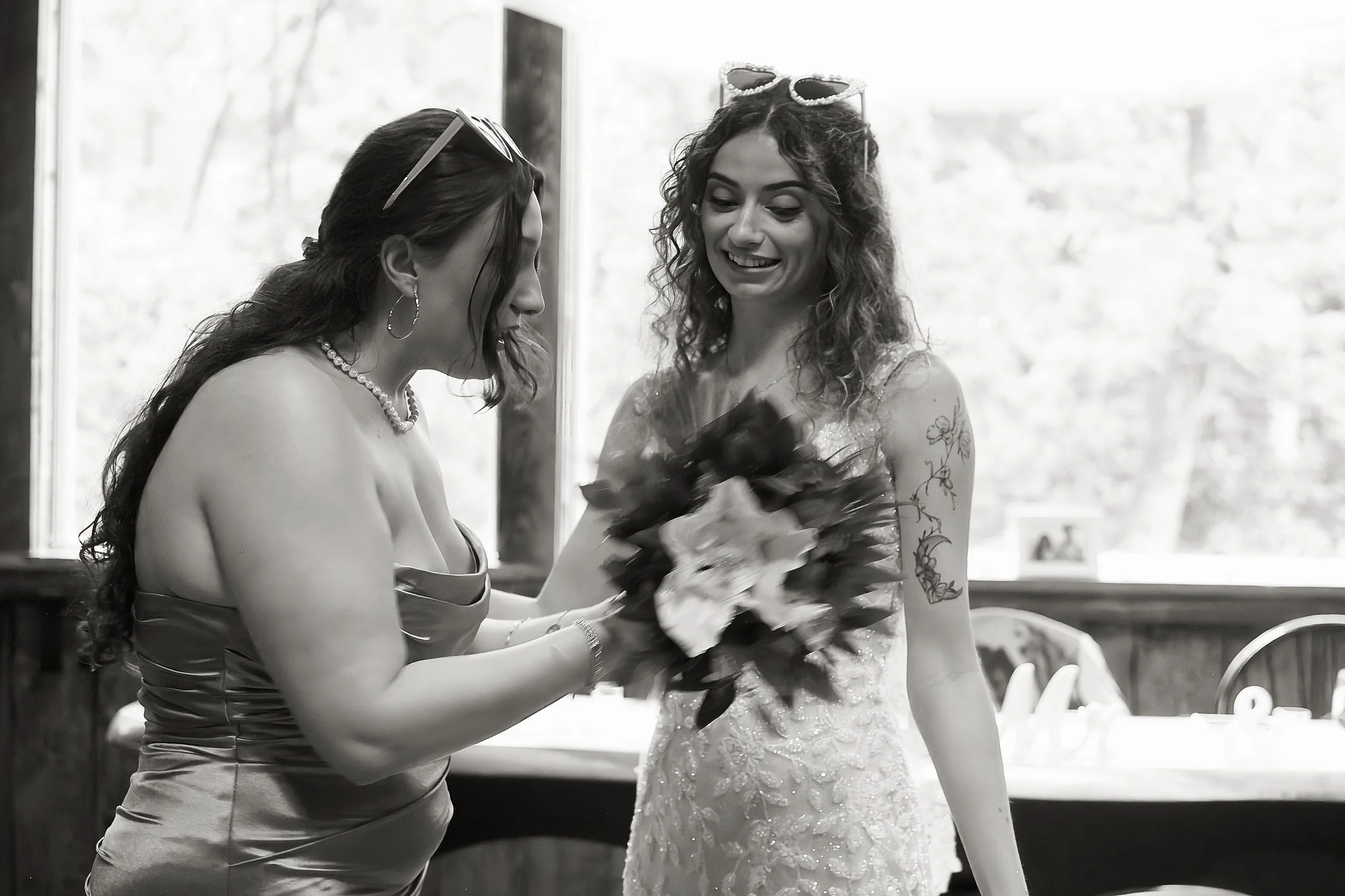 Two women in elegant dresses, one holding a bouquet, smiling and looking at each other in a bright room with large windows.
