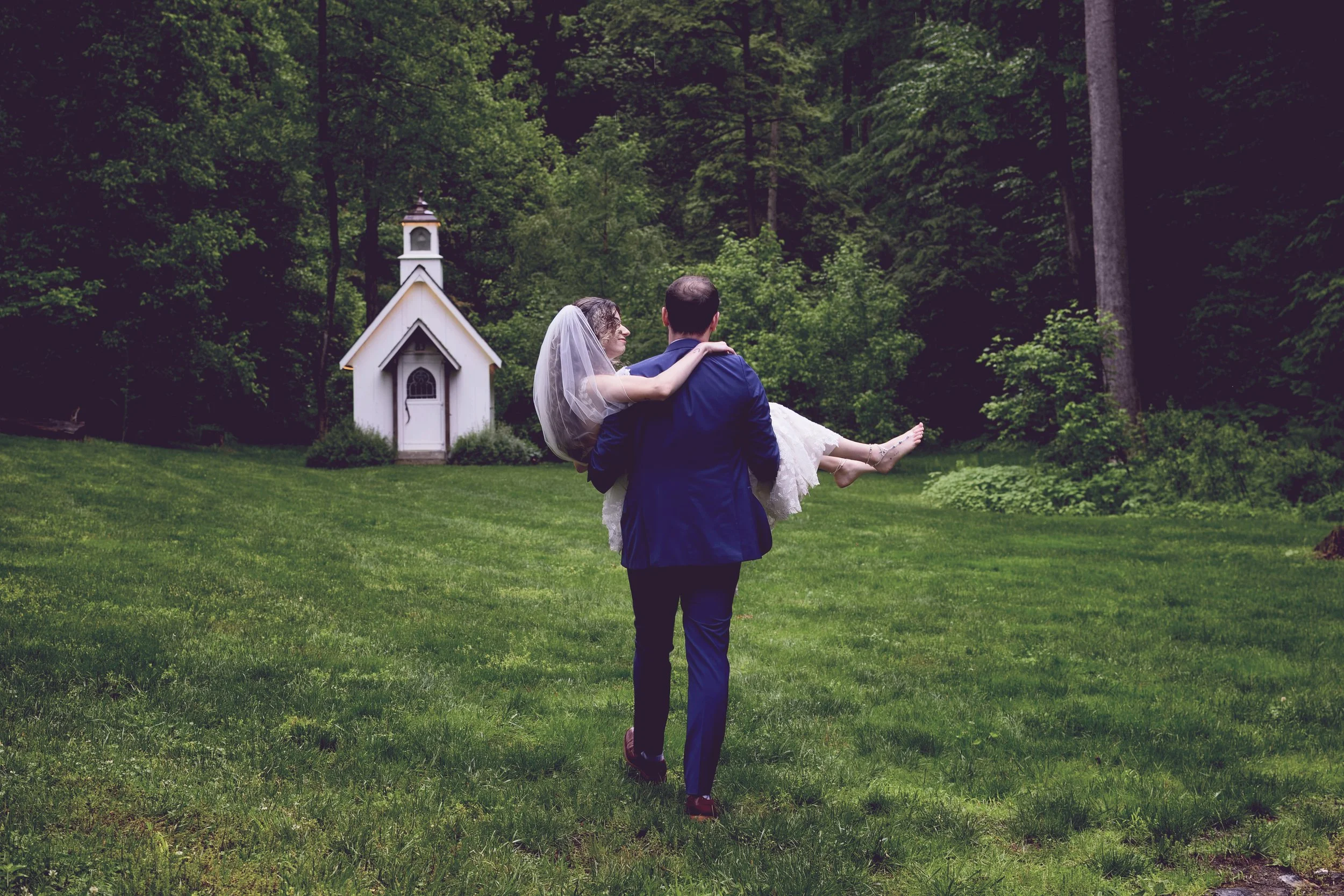 A groom in a blue suit carries a bride in a white wedding dress and veil through a green outdoor area with trees and a small white chapel in the background.