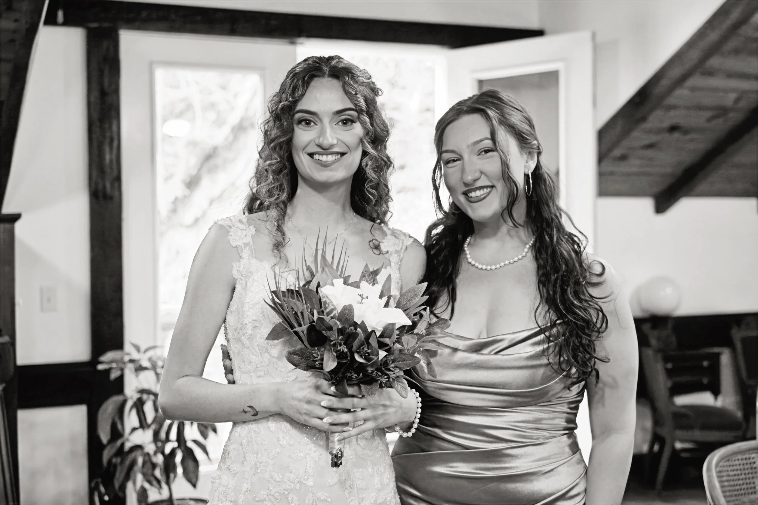 Two women smiling indoors, one dressed in a wedding gown holding a bouquet of flowers, and the other wearing a satin dress with pearl jewelry.