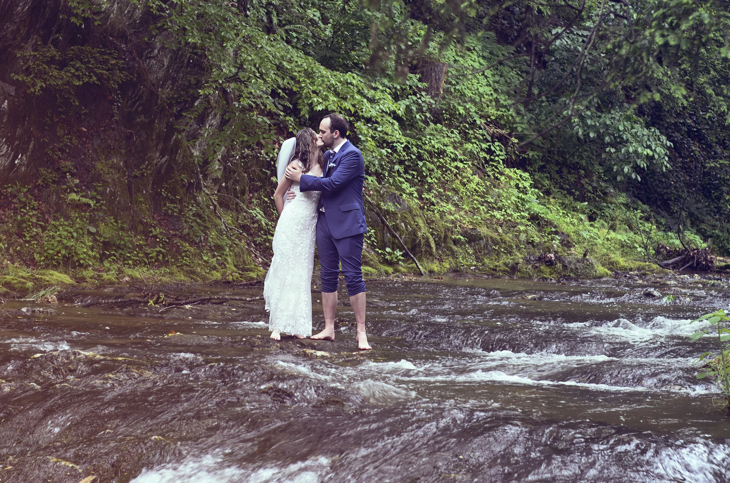A bride and groom standing barefoot in a shallow river, kissing surrounded by lush green trees and foliage.