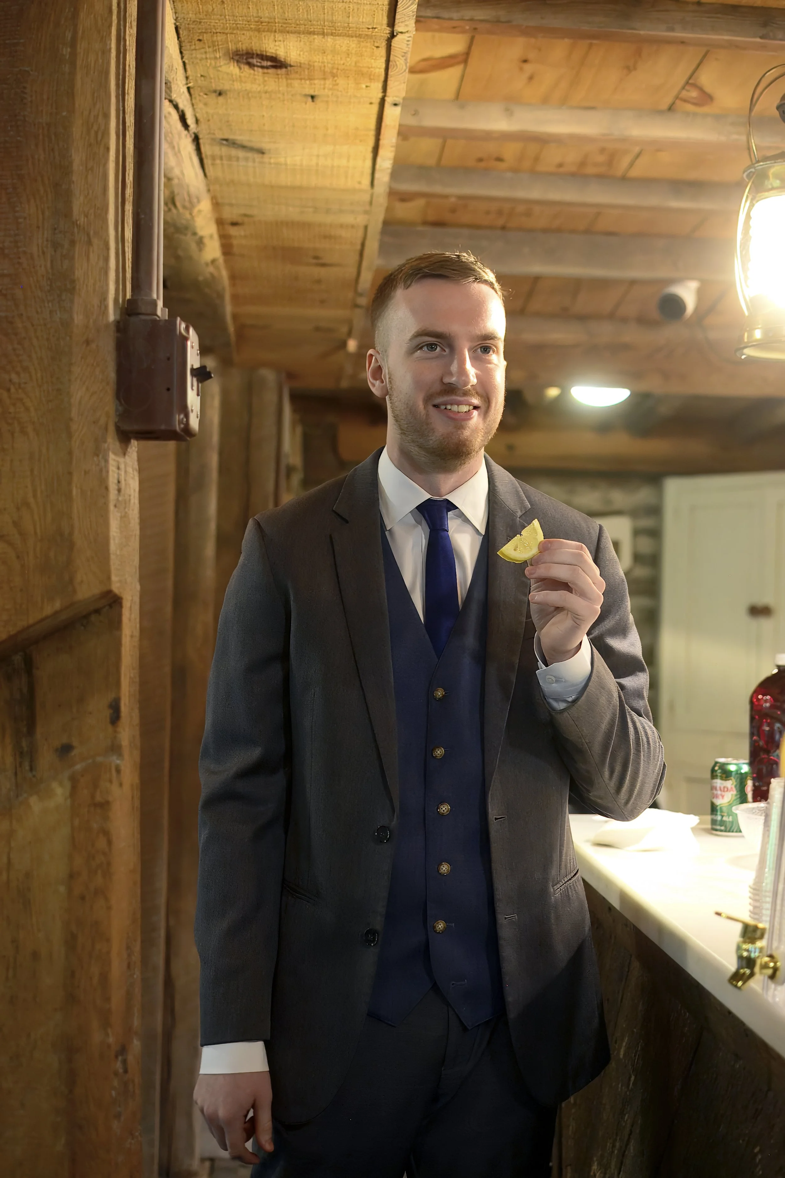 A man in a dark gray suit and blue tie holding a lemon wedge, standing in a wood-paneled room with warm lighting.