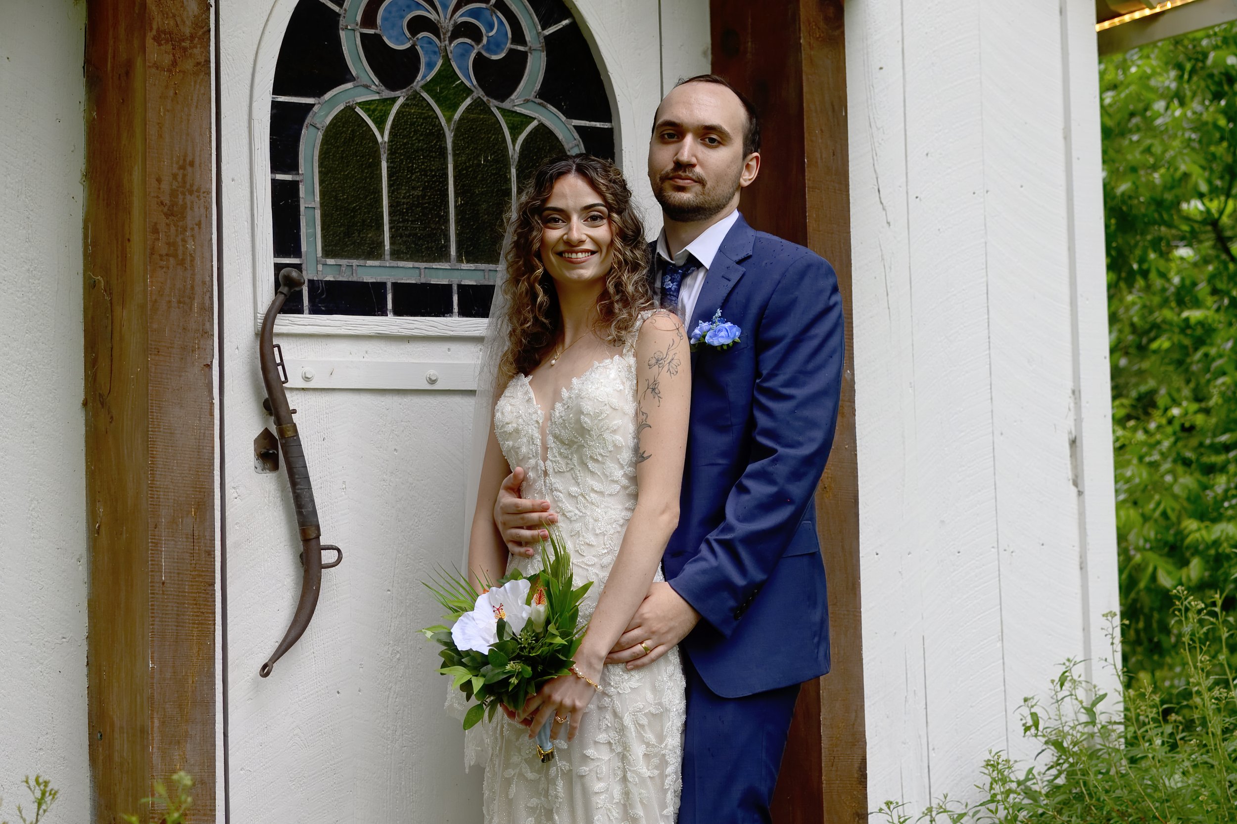 A newlywed couple poses outdoors against a rustic white wall with a stained glass window. The bride, with curly hair and tattoos on her arm, is smiling and holding a bouquet of white lilies, while the groom, with dark hair and beard, in a blue suit, 
