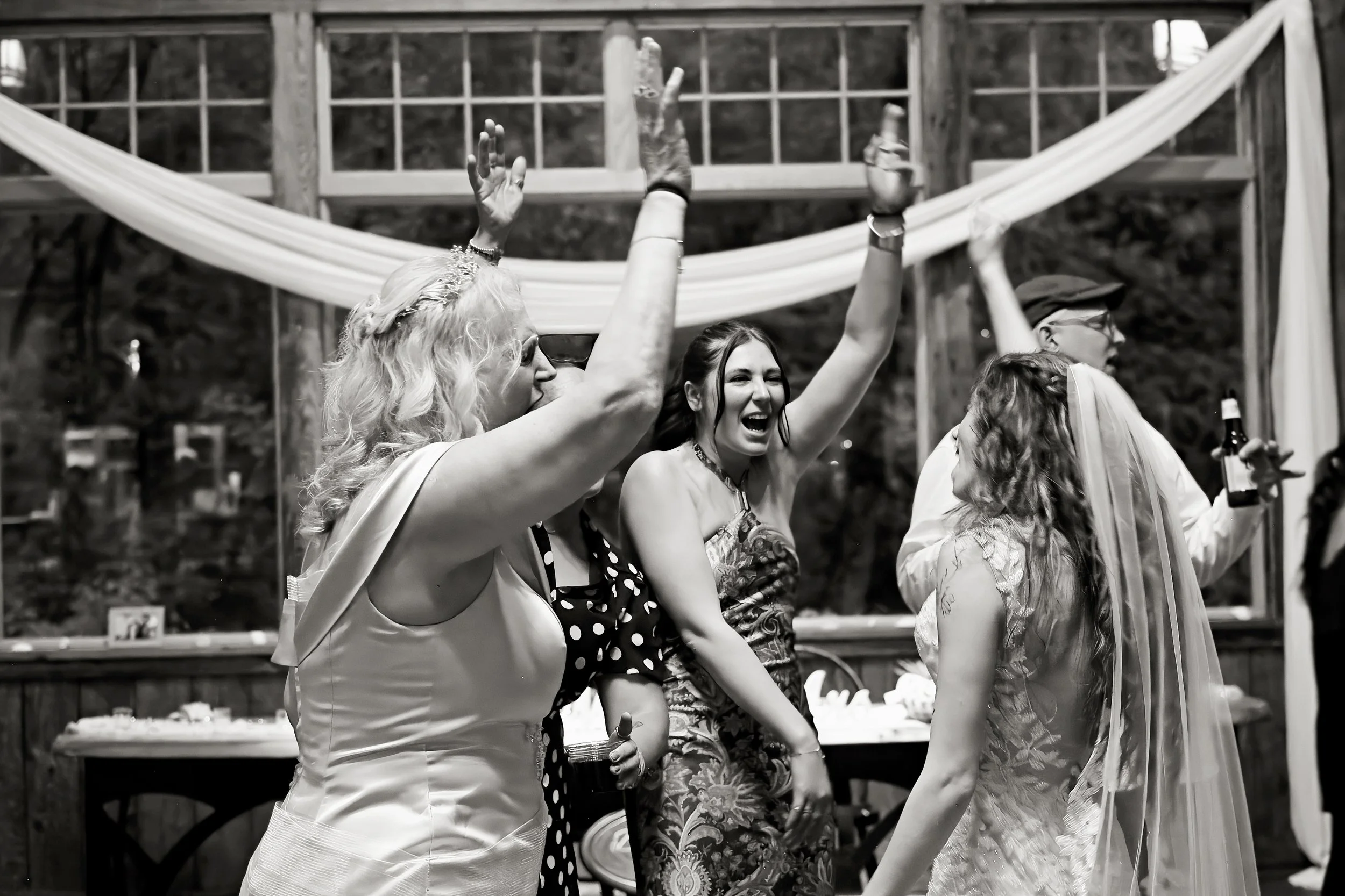 Group of women dancing and celebrating outdoors at what appears to be a wedding or celebration, with a wooden lattice and draped fabric in the background.