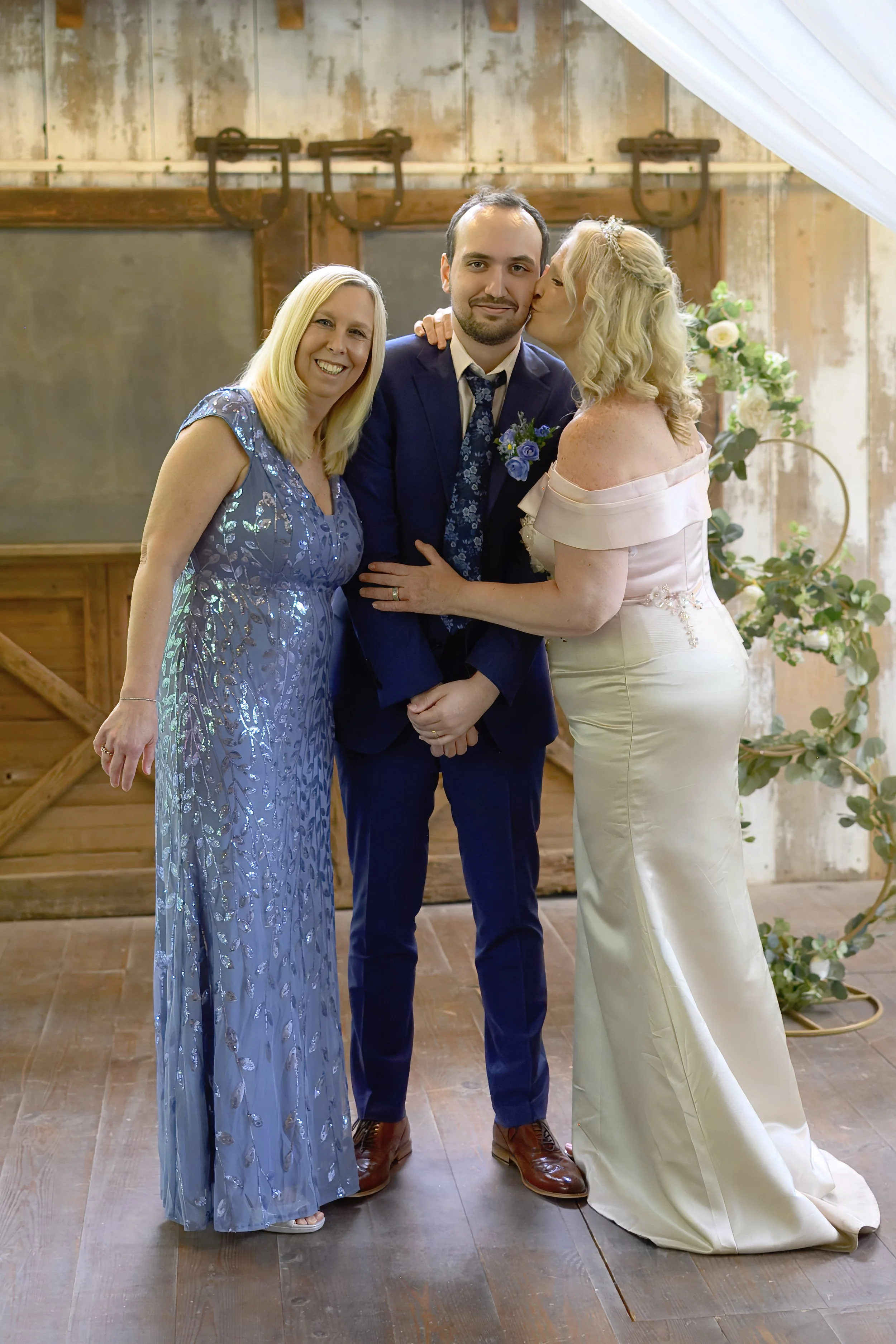 A wedding photo of a bride and groom with a woman. The bride is kissing the groom on the cheek, the groom is smiling, and the woman is smiling at the camera. The bride is wearing a satin off-shoulder wedding dress, the groom is in a navy suit with a 
