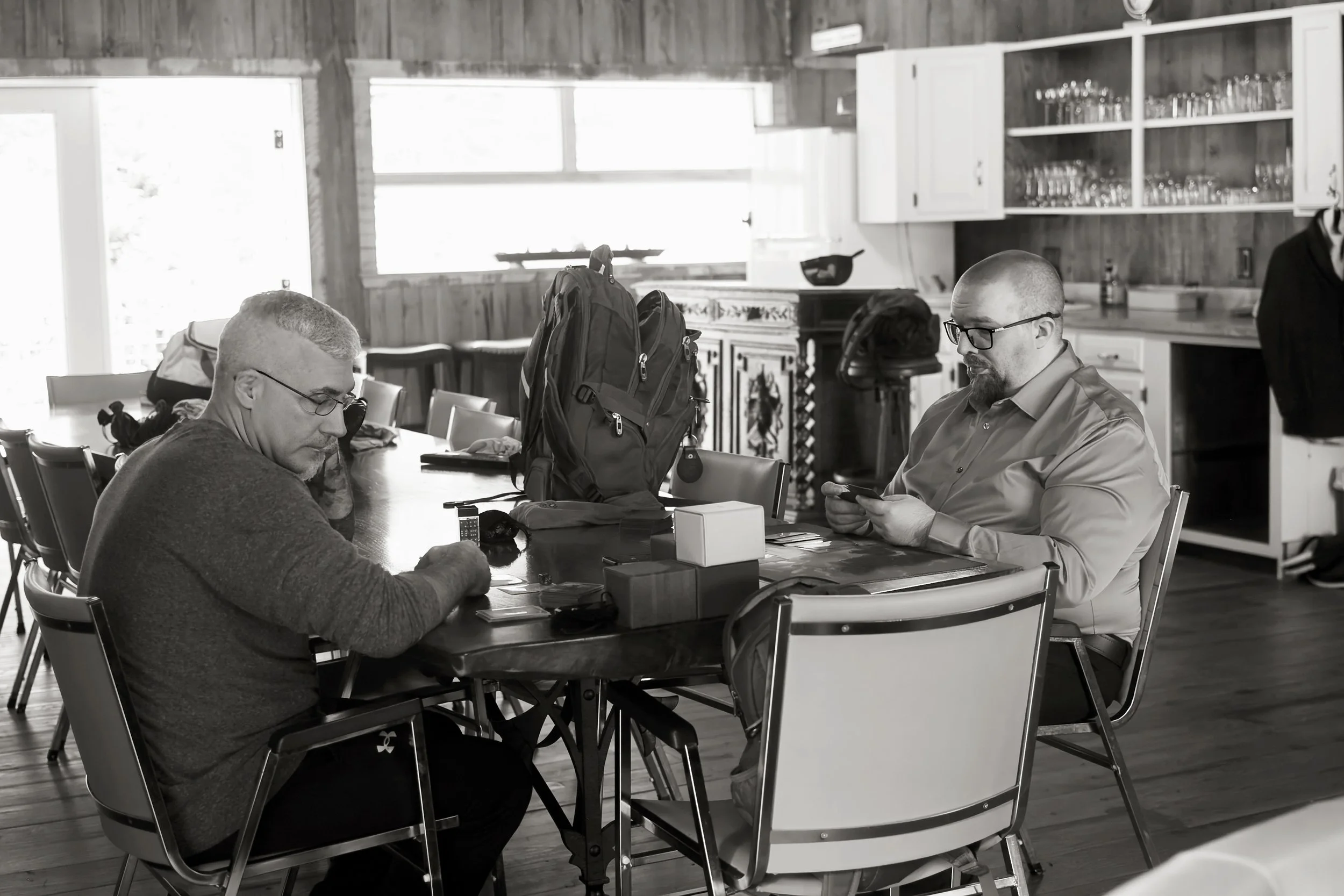 People sitting at a table with backpacks and electronic devices inside a rustic kitchen area with wooden walls, cabinets, and shelves of glasses.