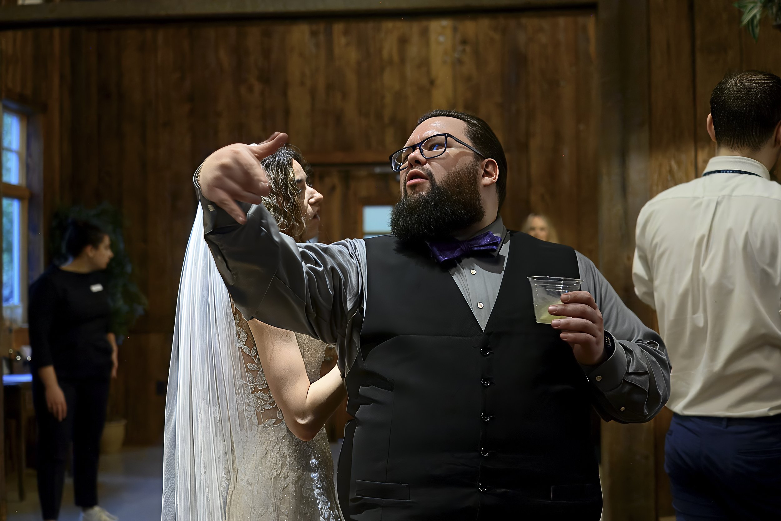 A man with a beard, glasses, and a bow tie dancing closely with a woman in a wedding dress at a reception. Other people are visible in the background in a wooden-paneled room.