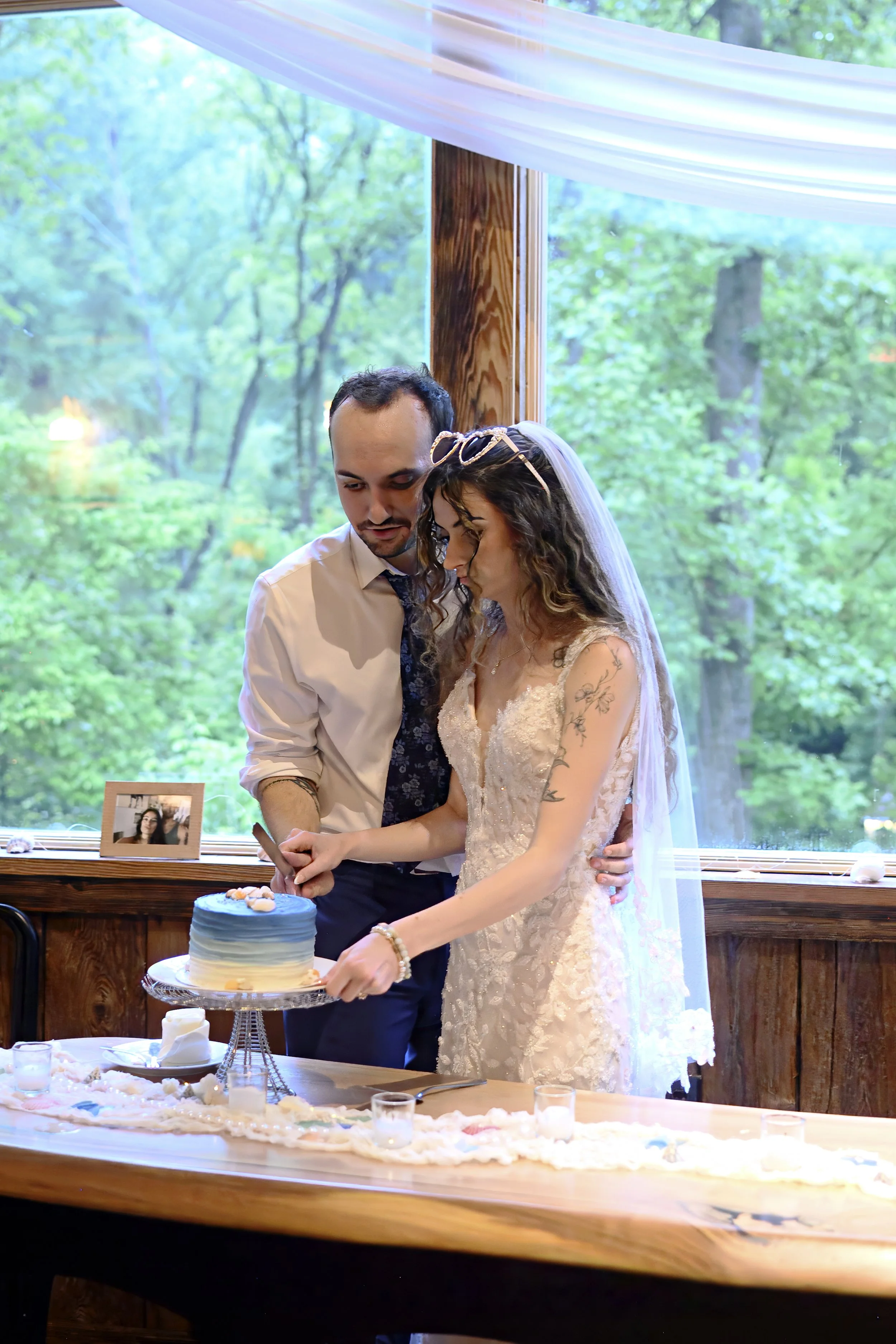 A bride and groom cutting a wedding cake indoors beside a large window with green trees outside, with a framed photograph and decorative tablecloth on the table.