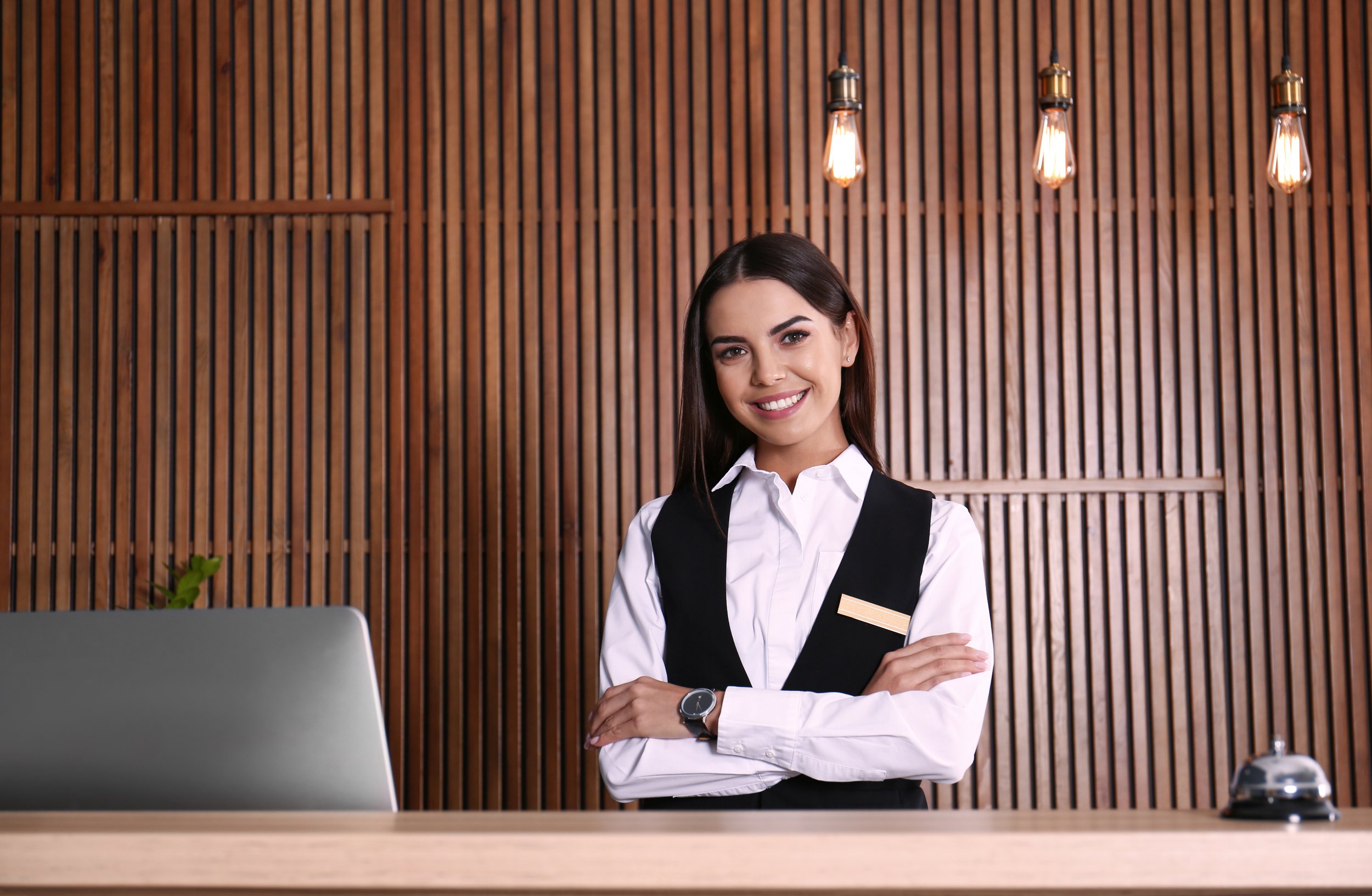 Smiling woman in a white shirt and black vest standing behind a reception desk, holding a small book or menu, in a modern interior with wooden wall panels and pendant lights.