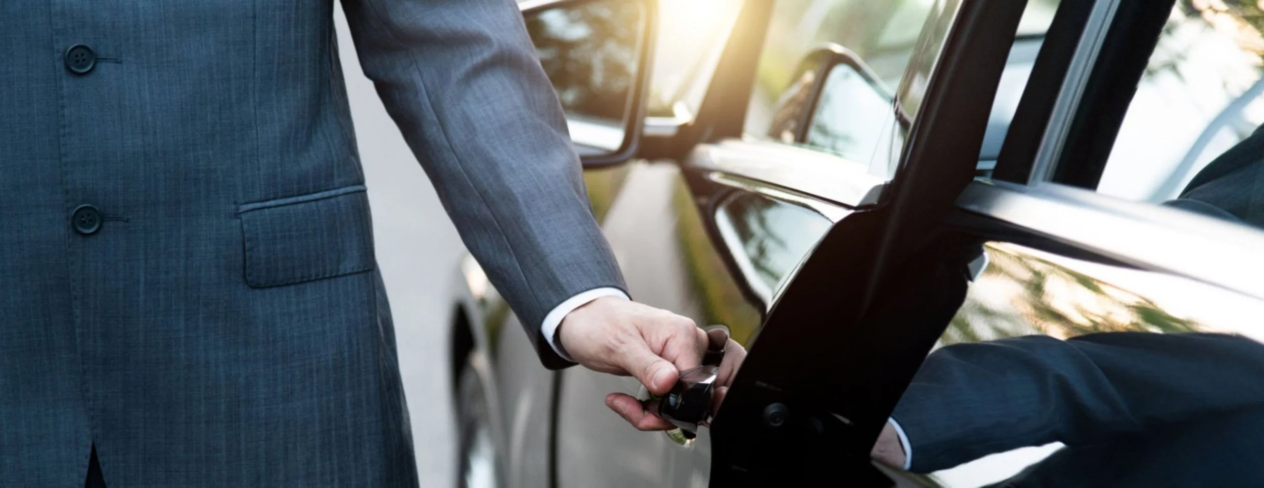 A person in a gray suit holding a car key near an open car door, with a touchscreen display inside the vehicle.