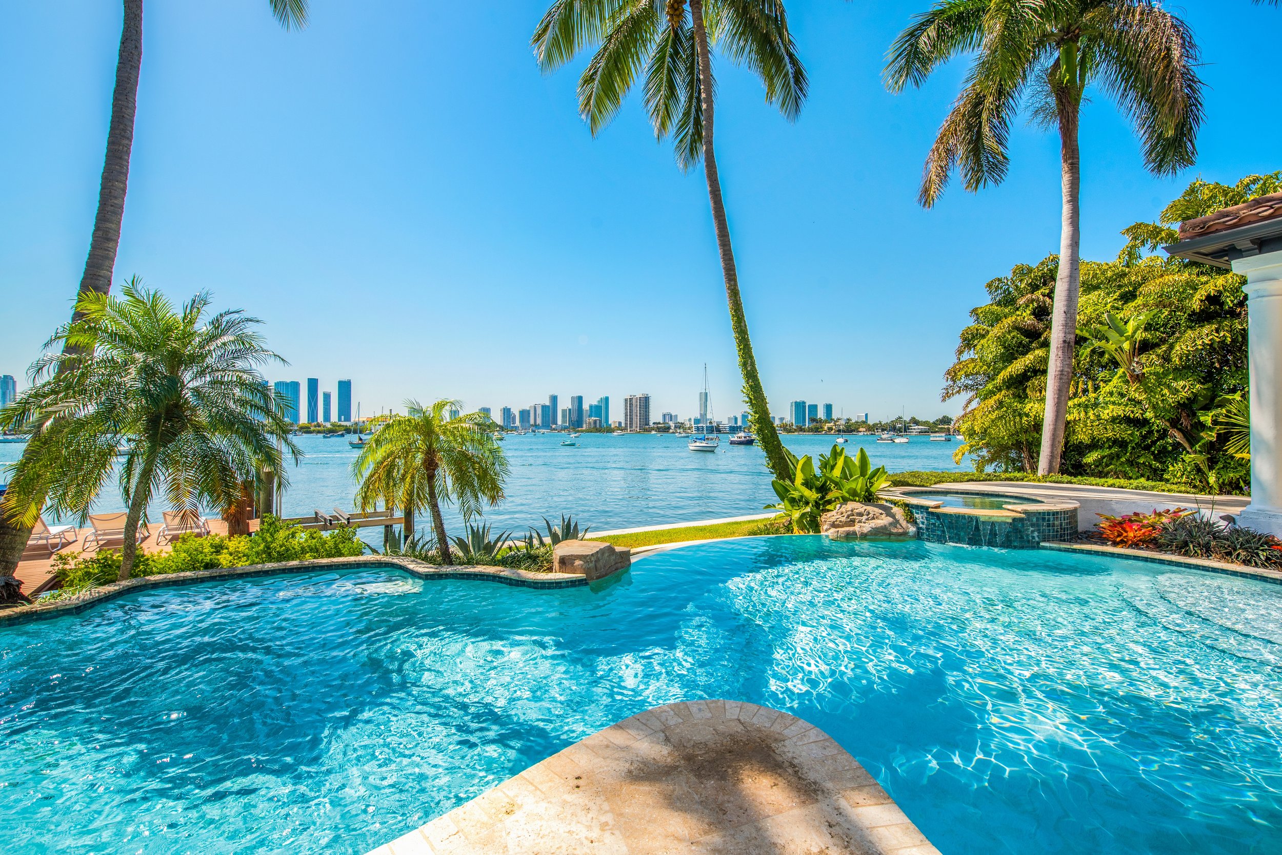 A luxurious outdoor swimming pool near the coast, with palm trees, lounge chairs, a hot tub, and a city skyline in the background on a sunny day.