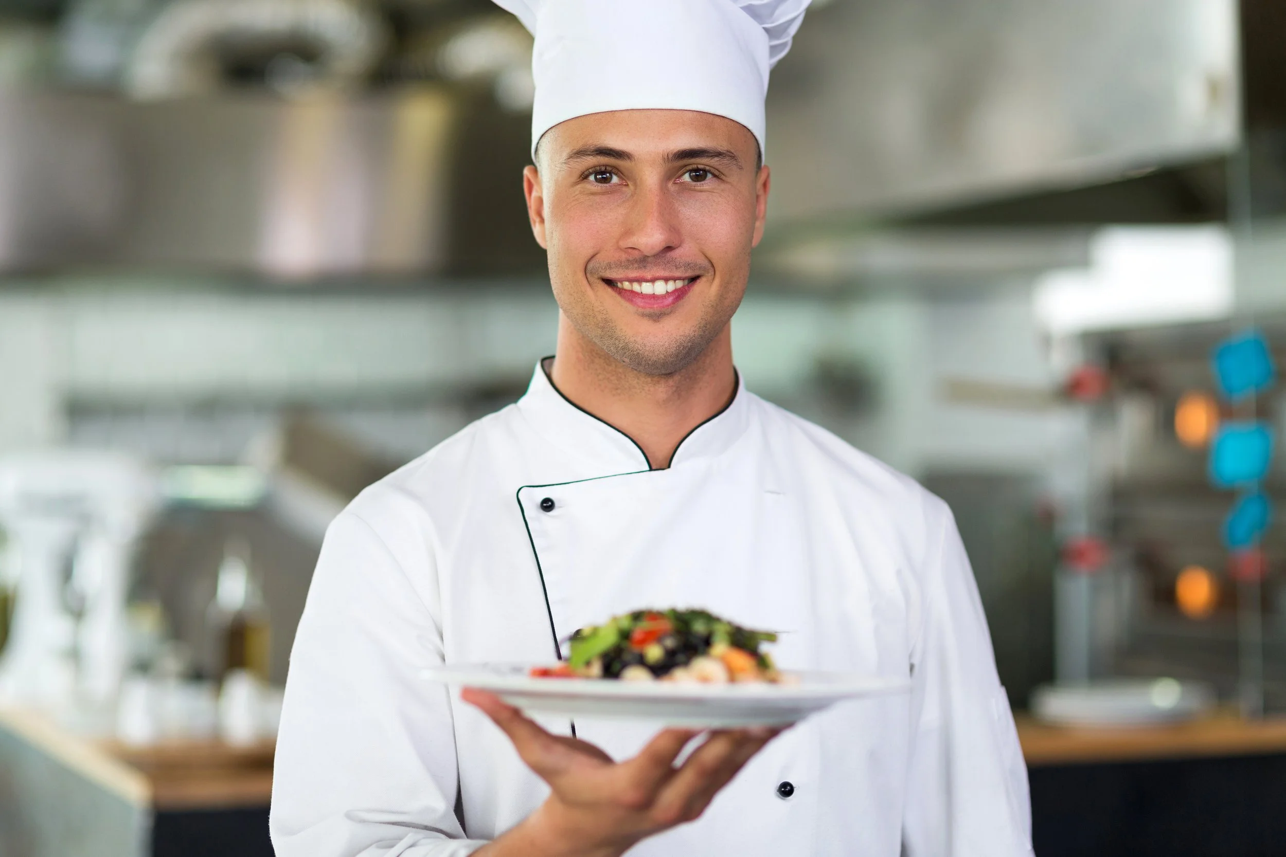 A smiling male chef with a white chef's hat and uniform holding a plate of salad in a commercial kitchen.
