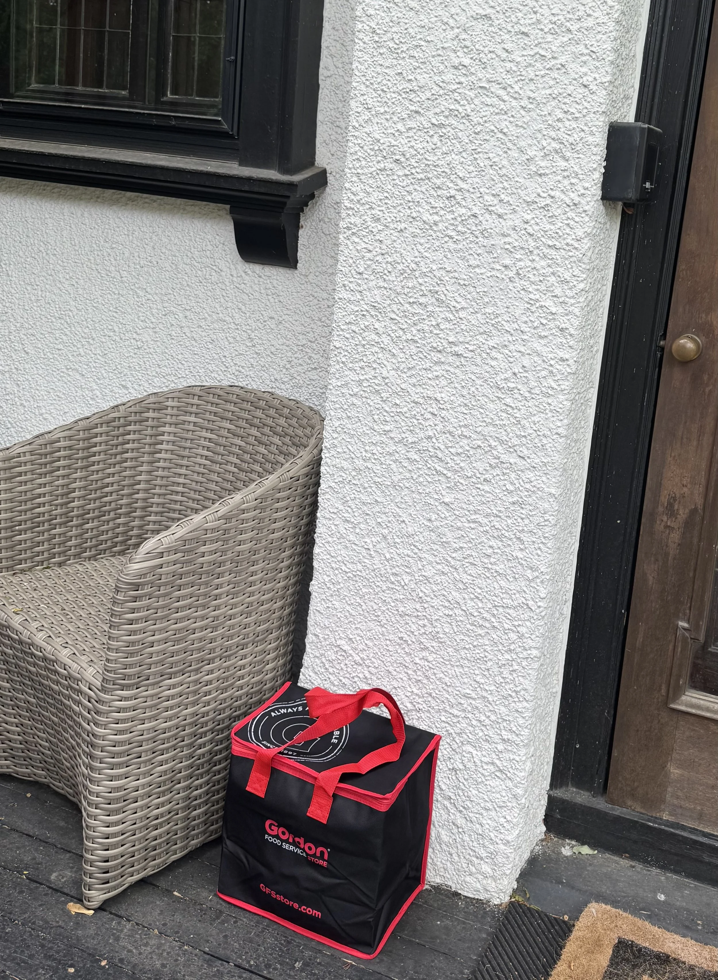 A black and red GFS store food delivery bag placed on the ground outside a house, next to a wicker chair, with a white textured wall and a black door frame in the background.