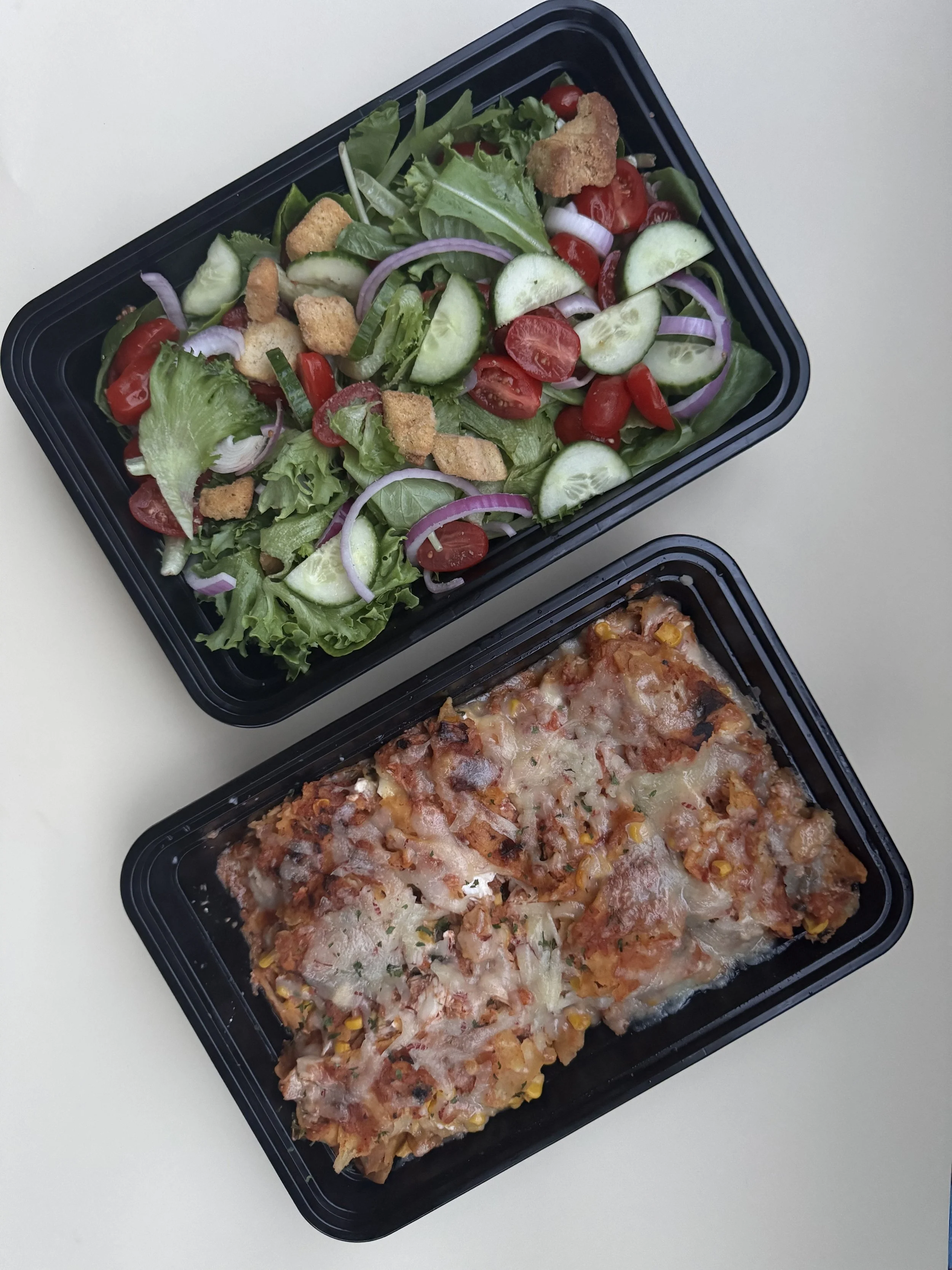 A black tray of vegetable salad with cherry tomatoes, cucumbers, red onions, lettuce, and croutons. Partially visible below is a black tray of baked pizza topped with cheese, corn, and diced tomatoes.