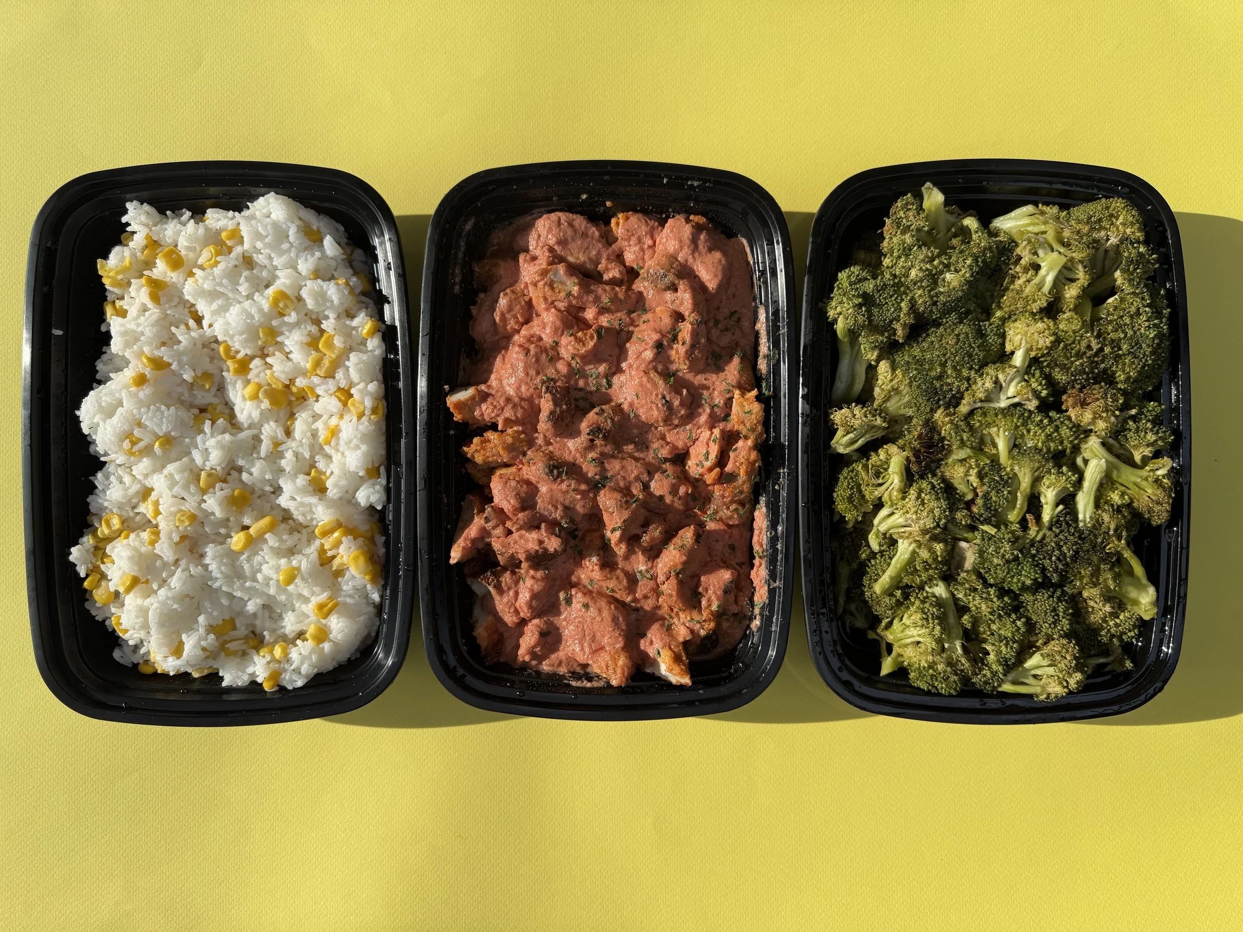 Three black food containers on a yellow surface, each filled with a different type of food: one with white rice mixed with corn, one with salmon in a creamy sauce, and one with broccoli florets.