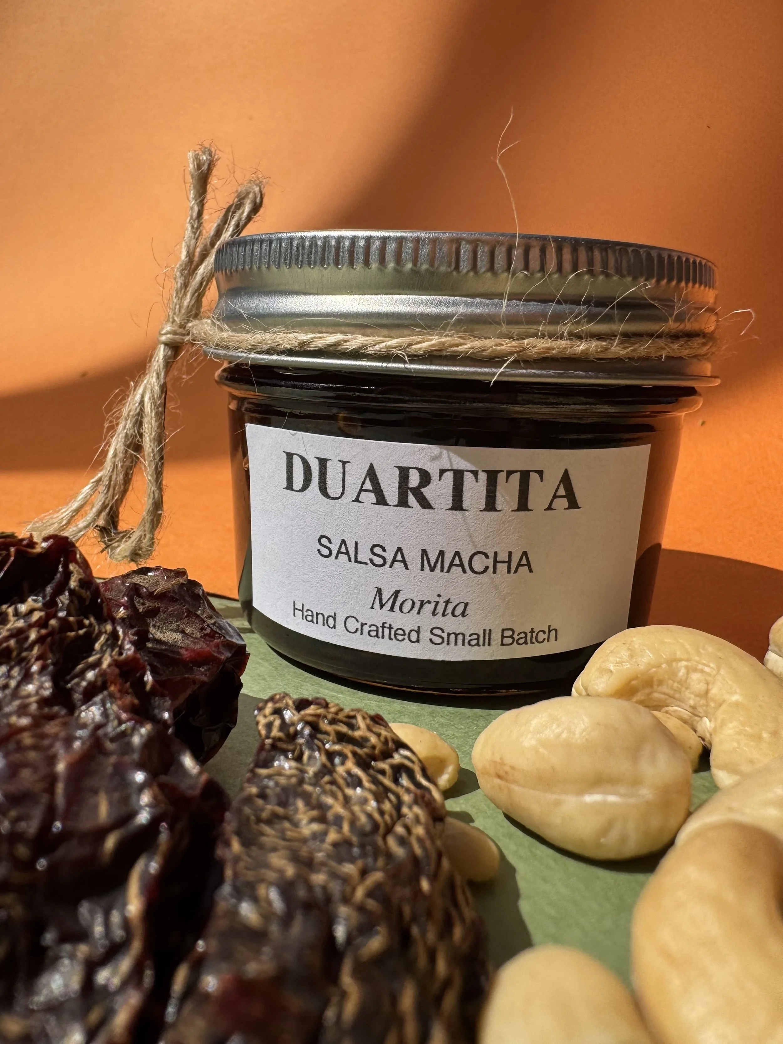 A jar labeled 'Duartita Salsa Macha' surrounded by dried chilies and yellow nuts against an orange background.