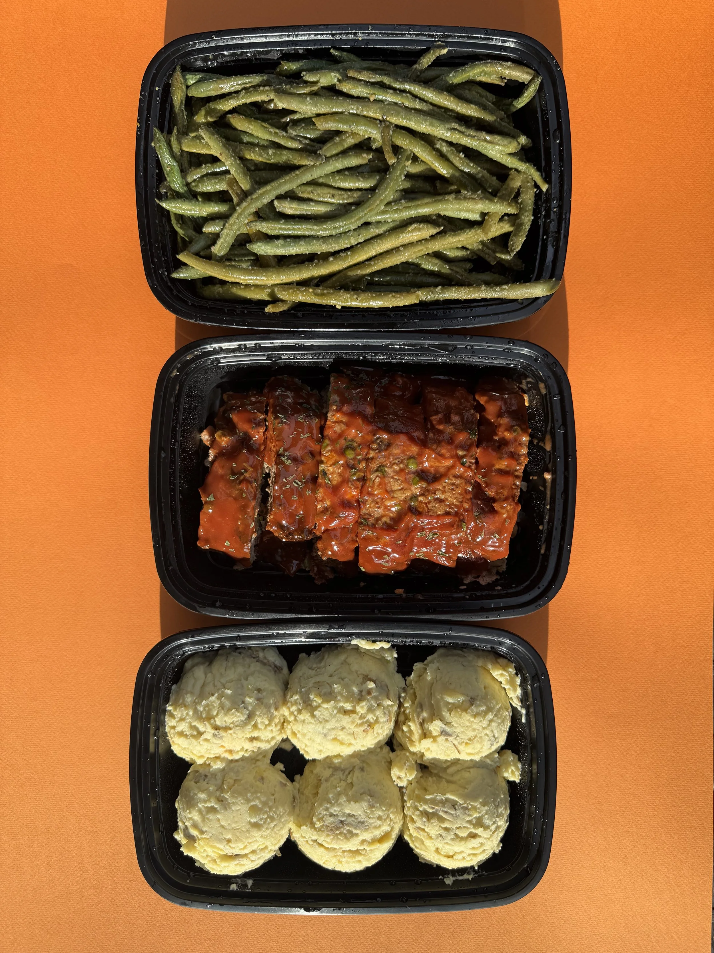 Three black plastic food containers stacked vertically on bright orange background, each containing a different dish. The top container has cooked green beans, the middle container has meatloaf with tomato sauce, and the bottom container has mashed p