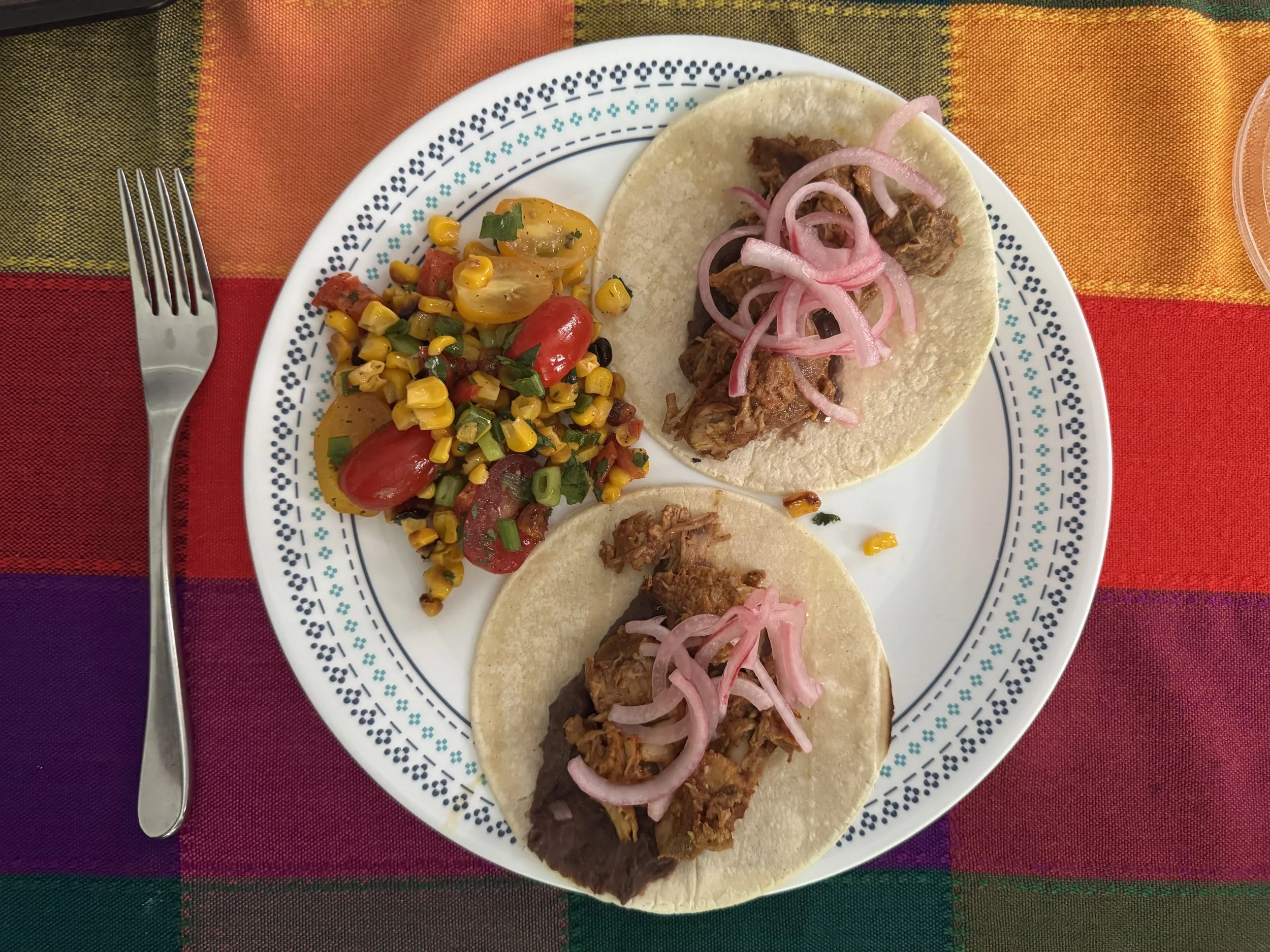 A white plate with blue and black decorative border, holding three tacos filled with shredded beef and topped with sliced onions, and a side of corn salad with cherry tomatoes, cilantro, and chopped green onions, on a colorful striped tablecloth.