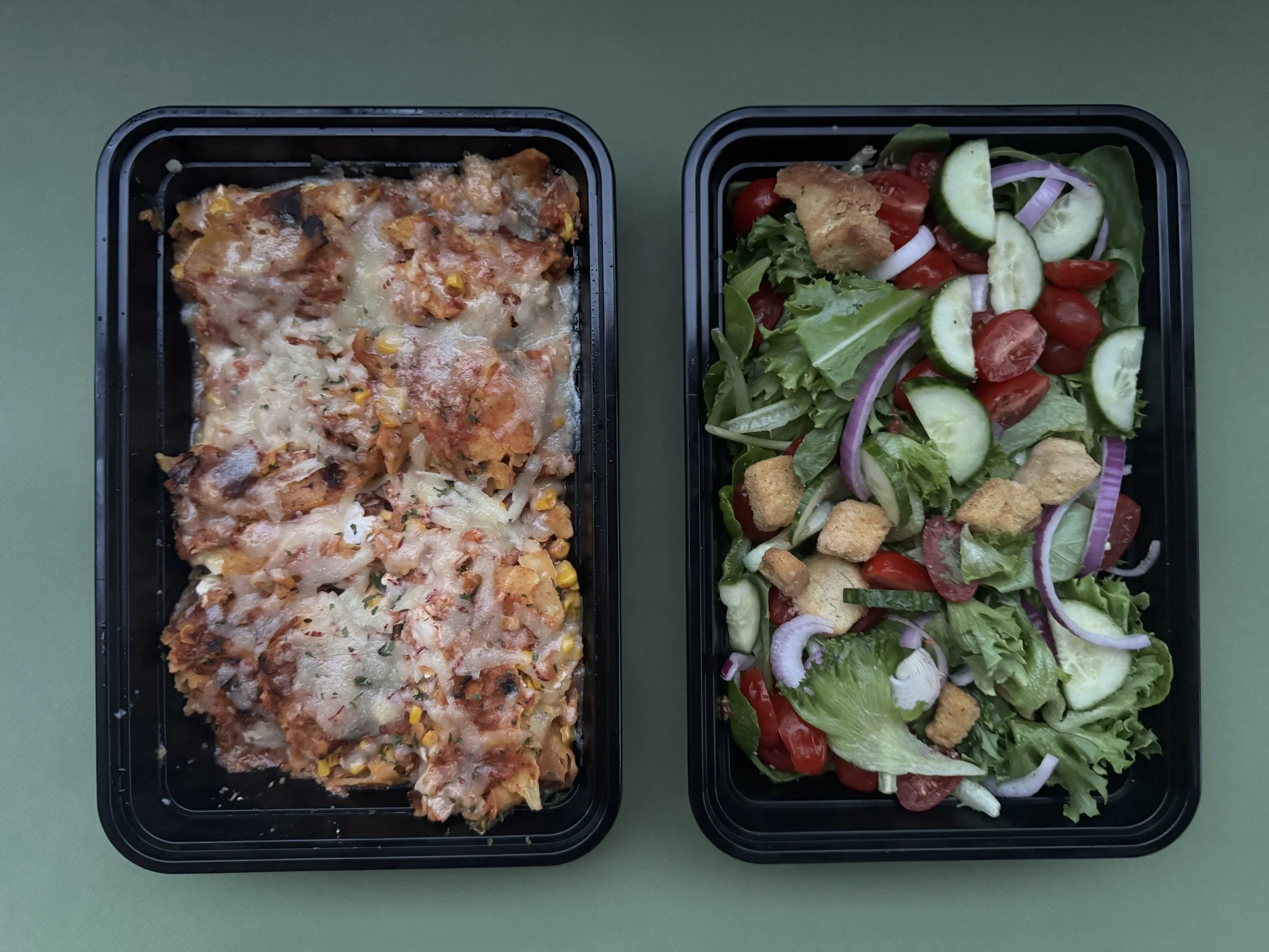 Two black plastic meal containers on a green surface. The container on the left holds a baked dish with cheese, corn, and tomato, with some melted cheese on top. The container on the right contains a fresh salad with cherry tomatoes, cucumber slices,