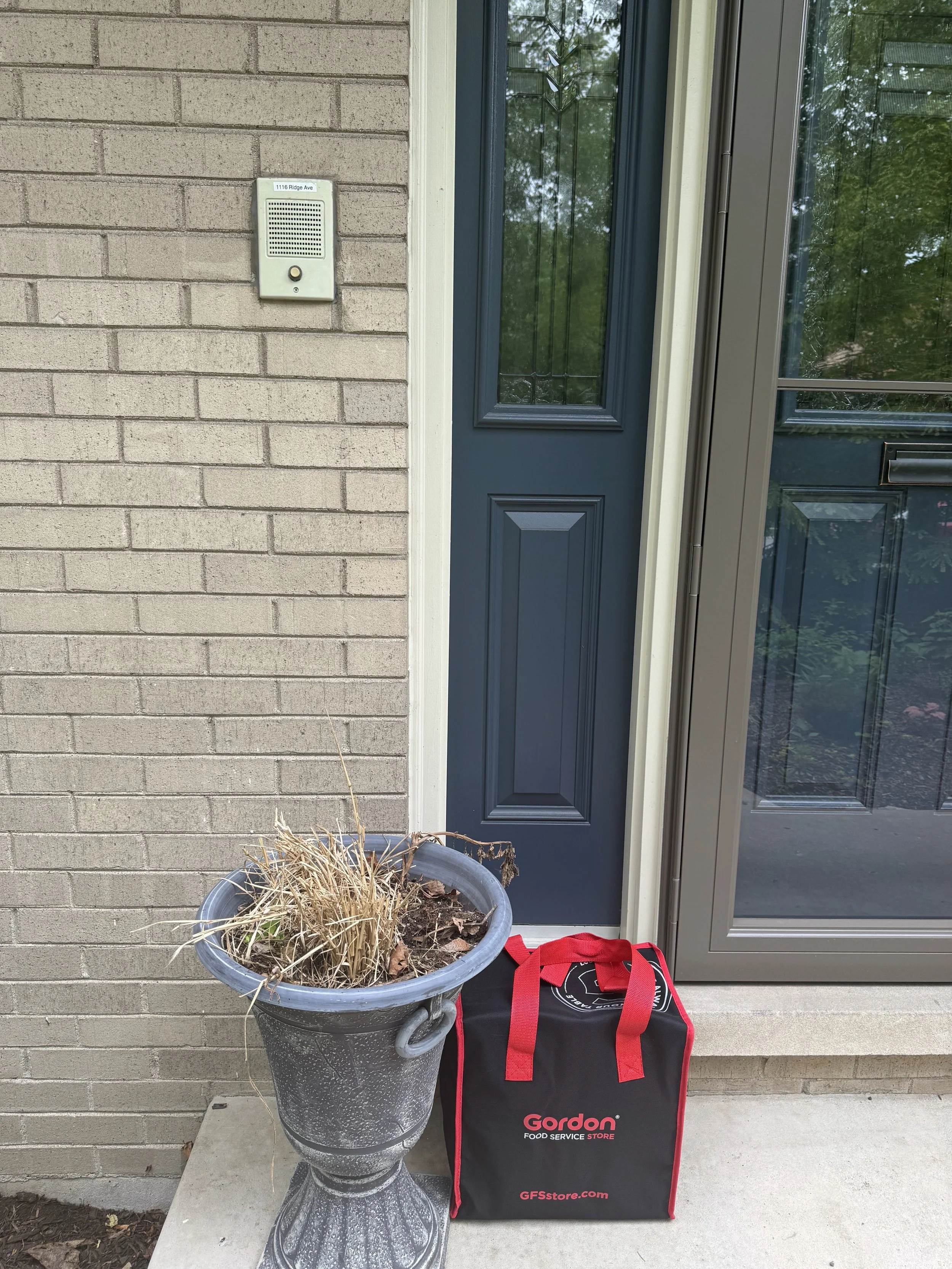 A large black foldable bag with red straps and the words 'Gordon Food Service Store' and 'GFSstore.com' written on it, placed next to a tall stone planter with dried plant remains in front of a blue door with glass panels, and a beige brick wall with