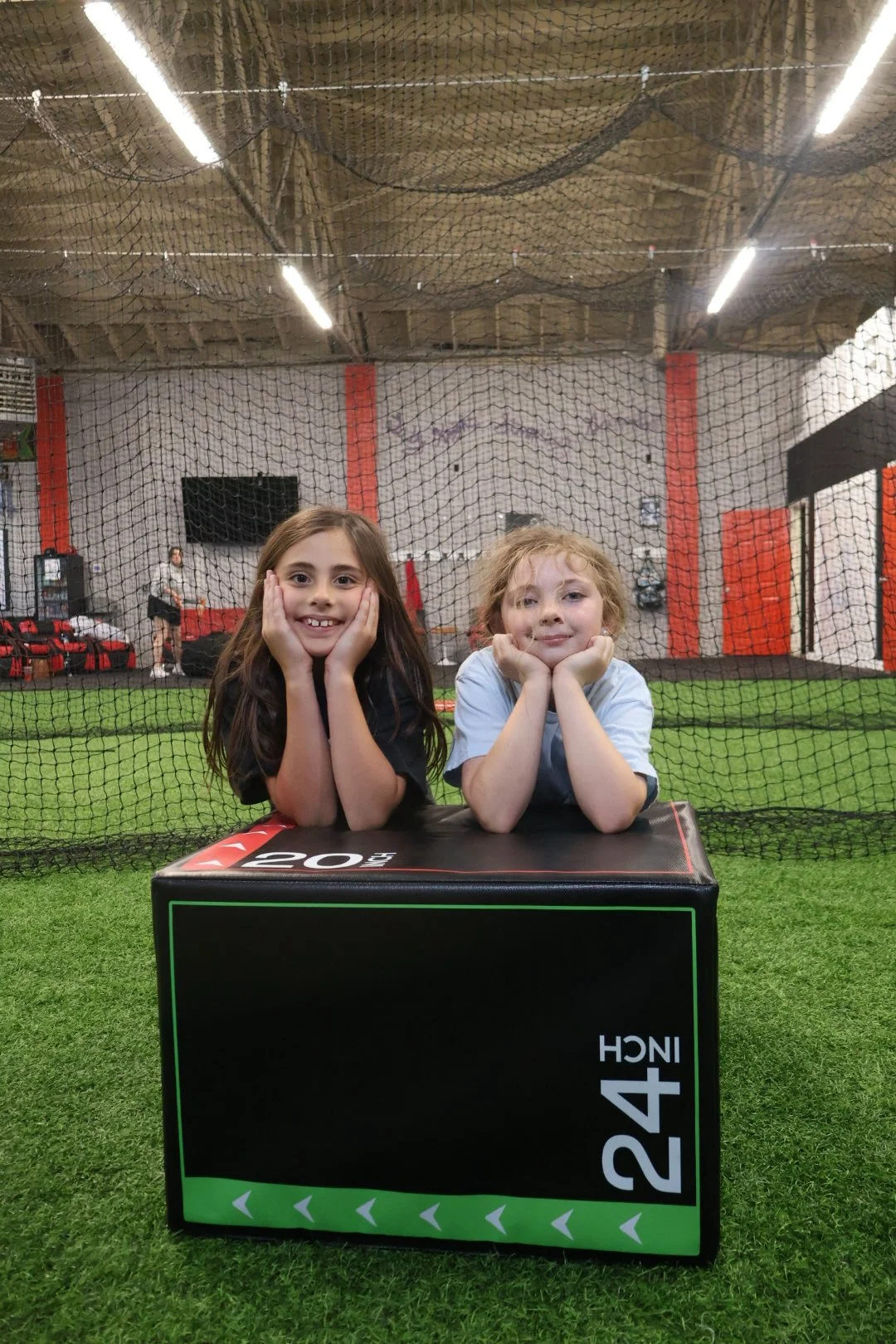 Two children lying on a black plyometric box with a 24-inch label, inside a sports facility with artificial grass, a netted area, and a TV screen in the background.