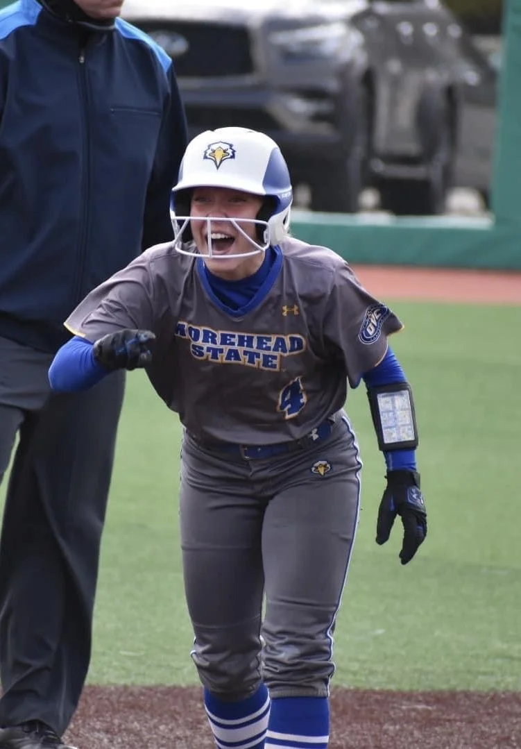 A female softball player in a grey and blue uniform celebrating on the field, wearing a helmet with a logo, and standing next to an adult, on a softball field.