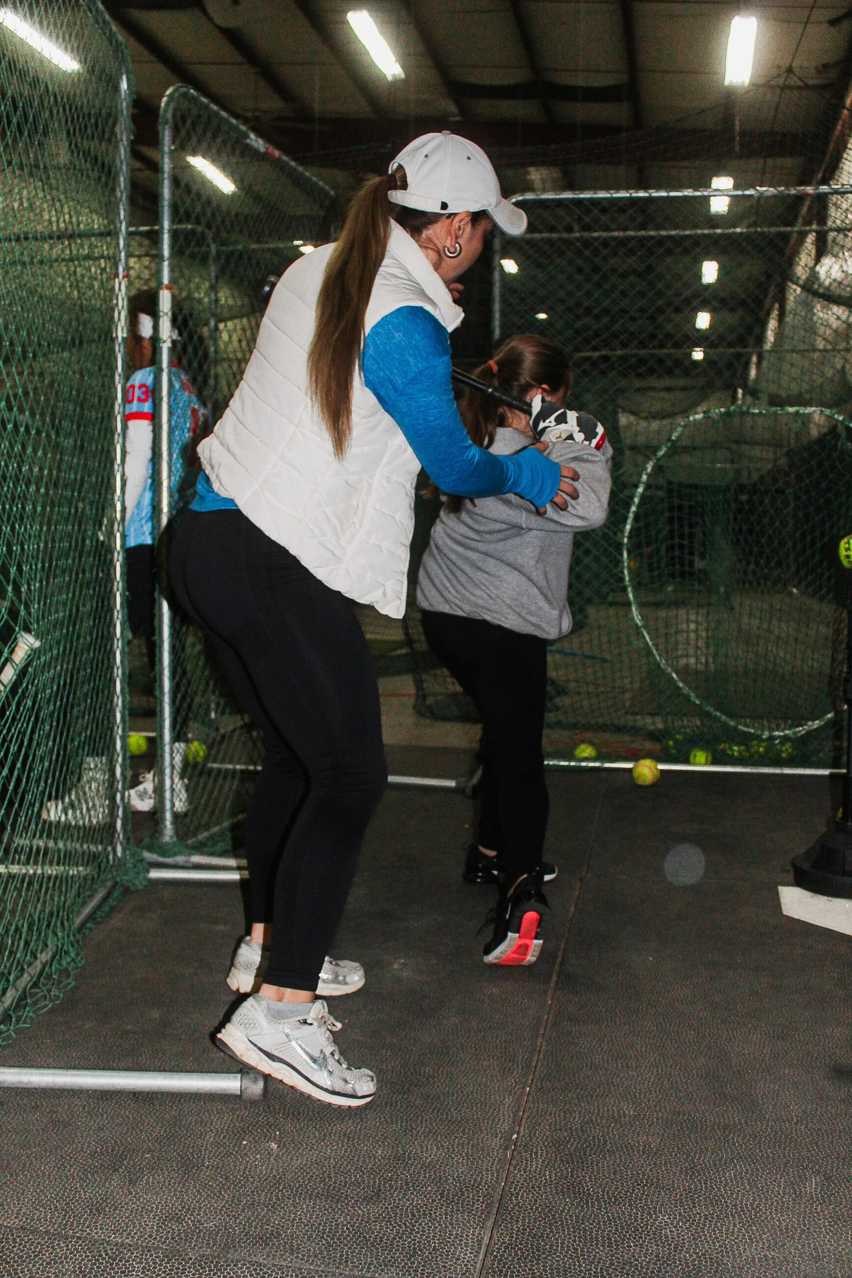 An adult woman is teaching a young girl how to play tennis in an indoor tennis court, with tennis balls and a net visible in the background.