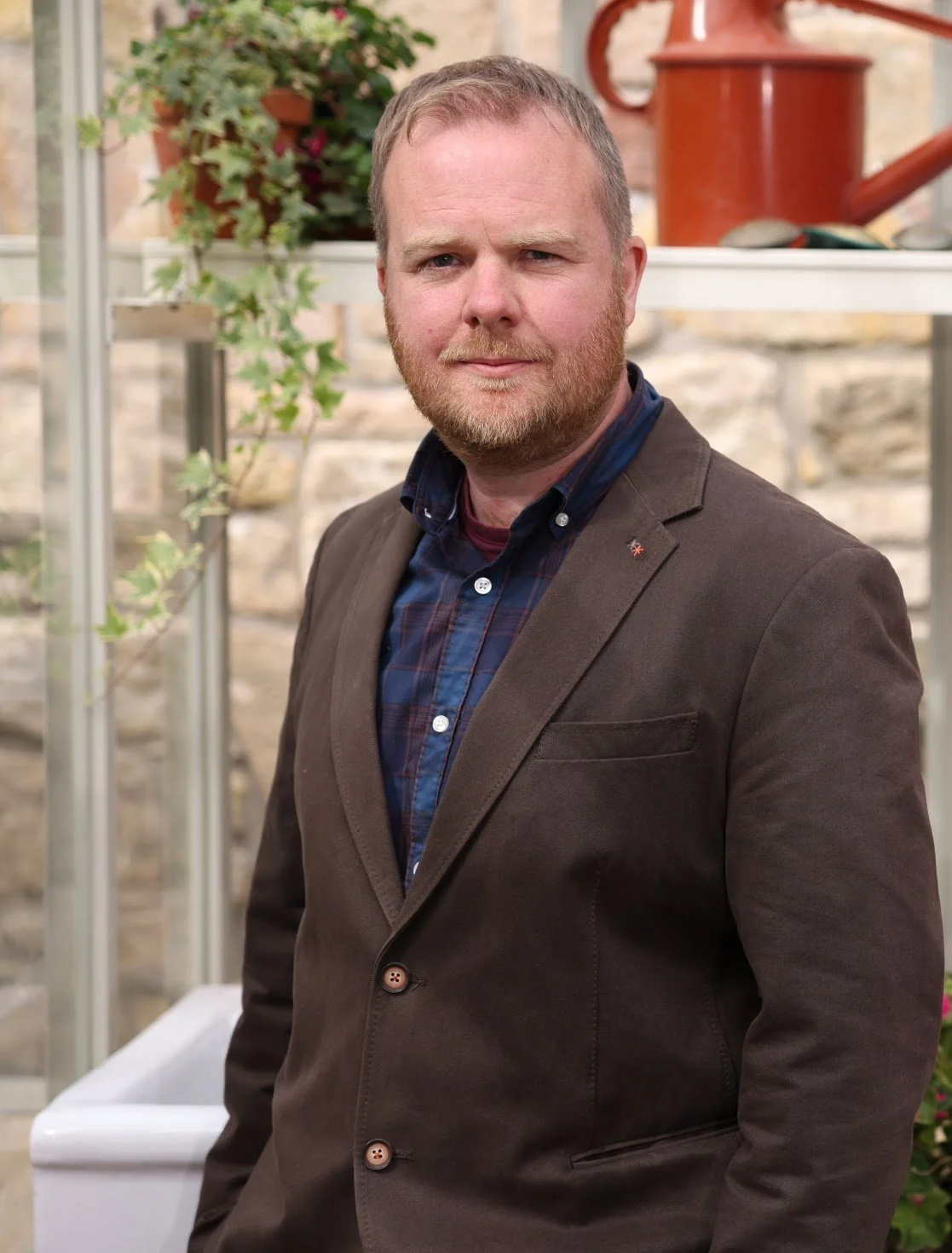 Portrait of a man with a beard and mustache, wearing a brown blazer over a blue plaid shirt, standing outdoors in front of a greenhouse with plants.
