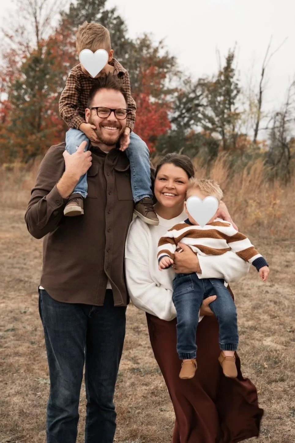 a mom in a field holding her son and standing next to her husband who is carrying their son on his shoulders