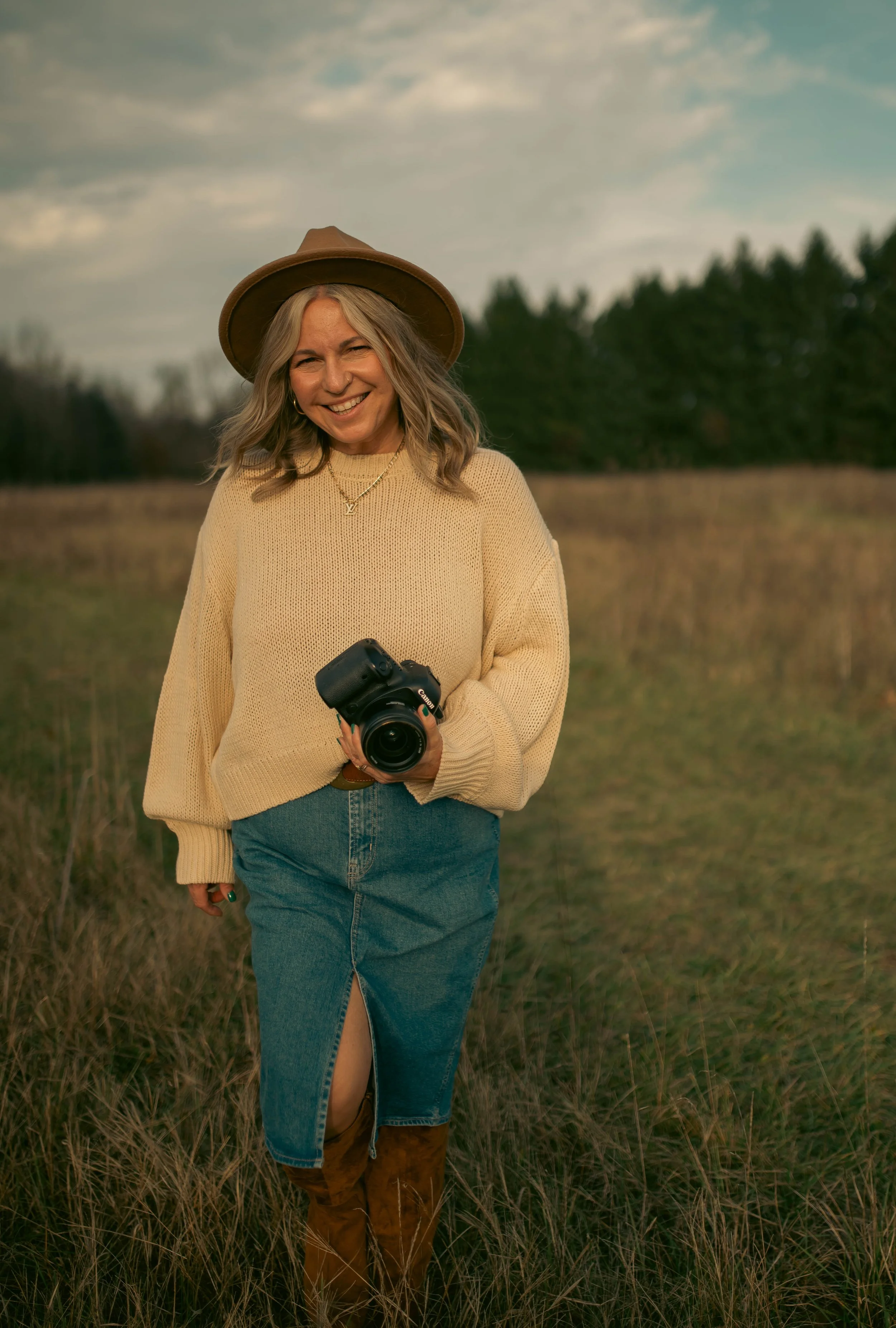 a woman smiling and holding a camera in a field and wearing a hat