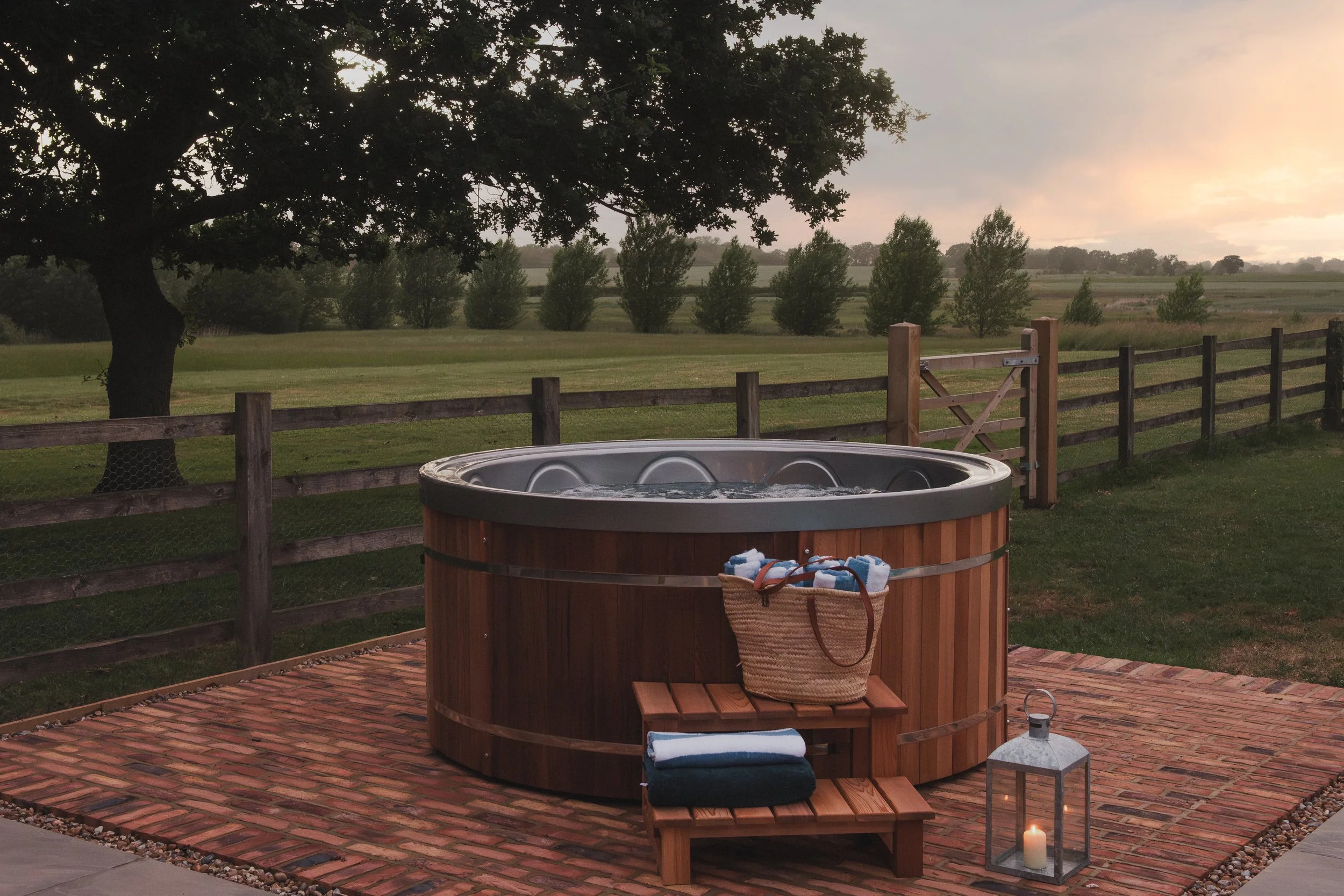 A backyard scene at sunset showing a hot tub on a brick patio, a wooden bench with folded towels, a lantern with a candle, a fence, and a landscape with trees and fields under a cloudy sky.