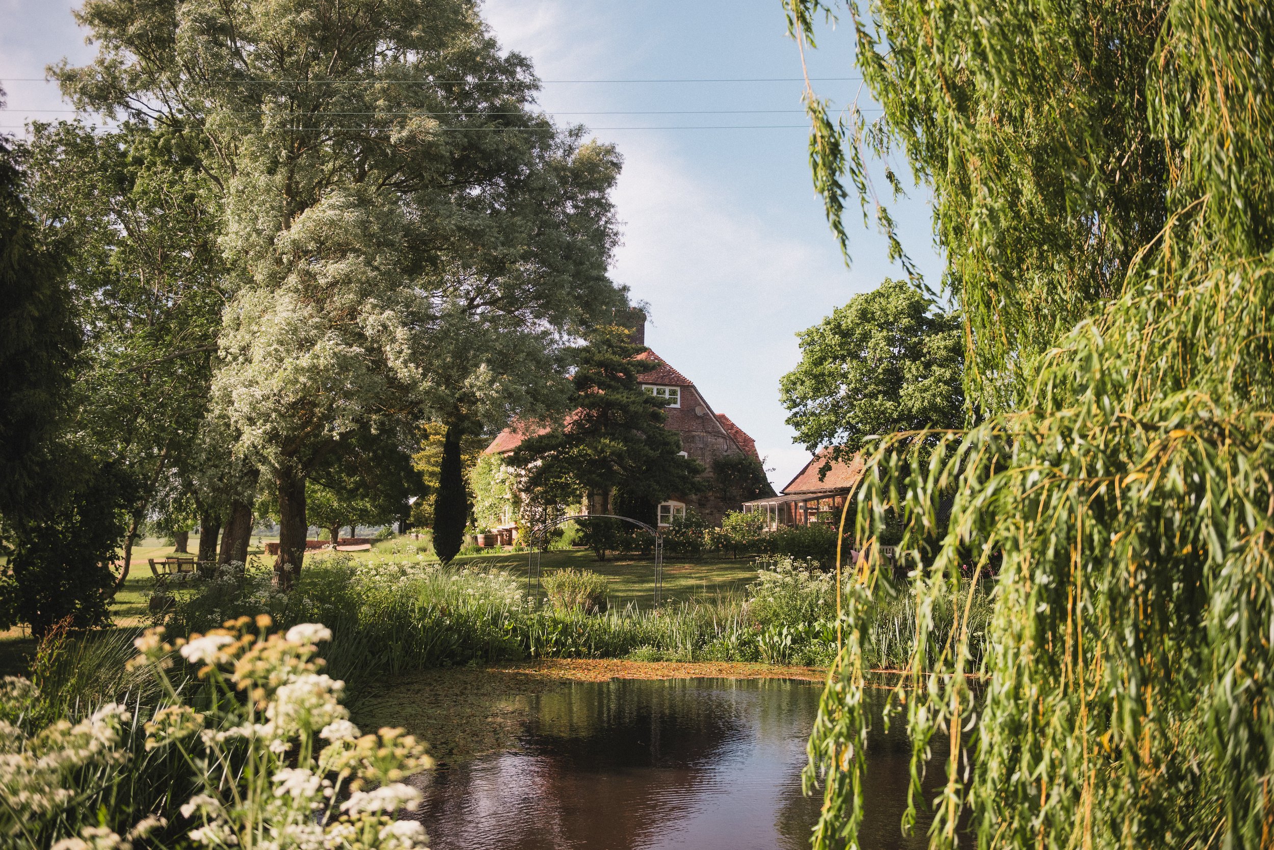 A peaceful garden scene with a small pond, tall trees, flowering bushes, and a red-roofed house in the background.