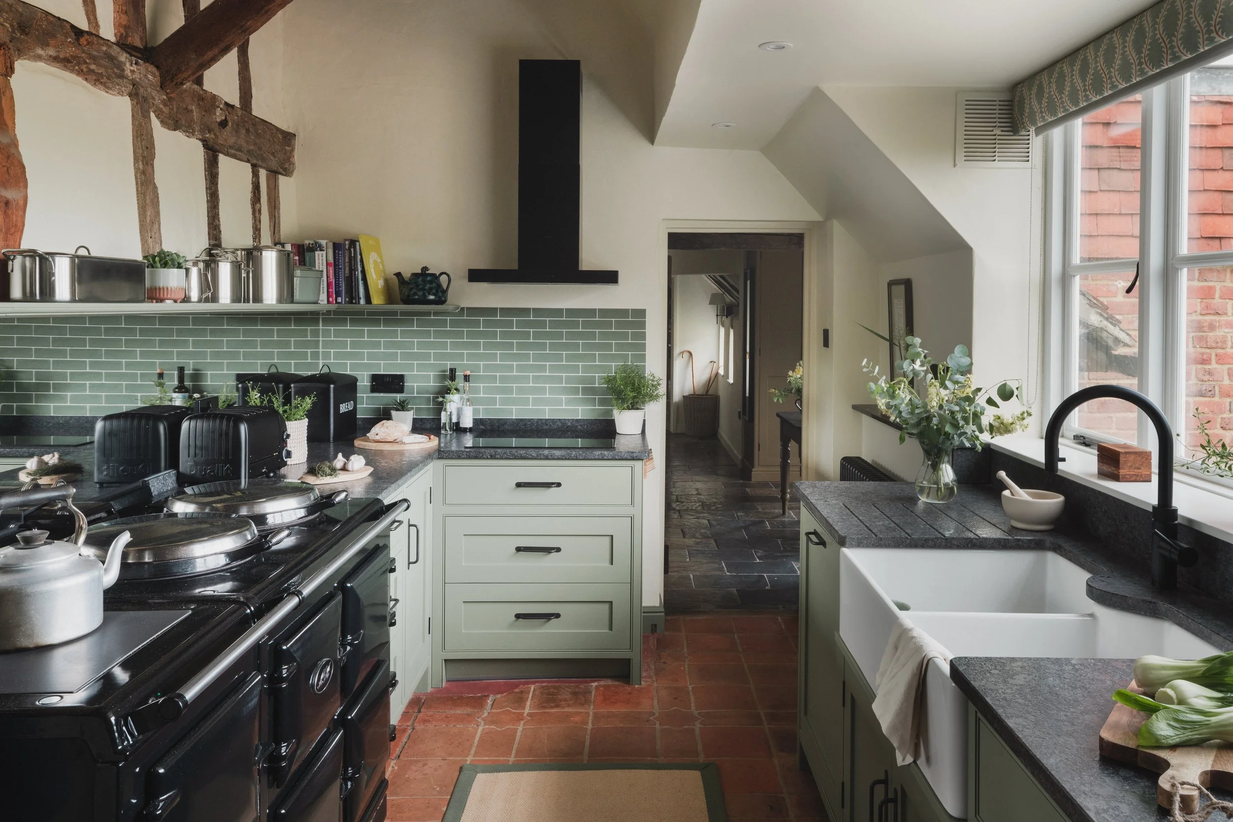 A cozy kitchen with green cabinets, black countertops, and a black stove. There are plants and kitchen utensils on the counter, and a window with a patterned curtain letting in natural light. The wall has a green tile backsplash and there are wooden 