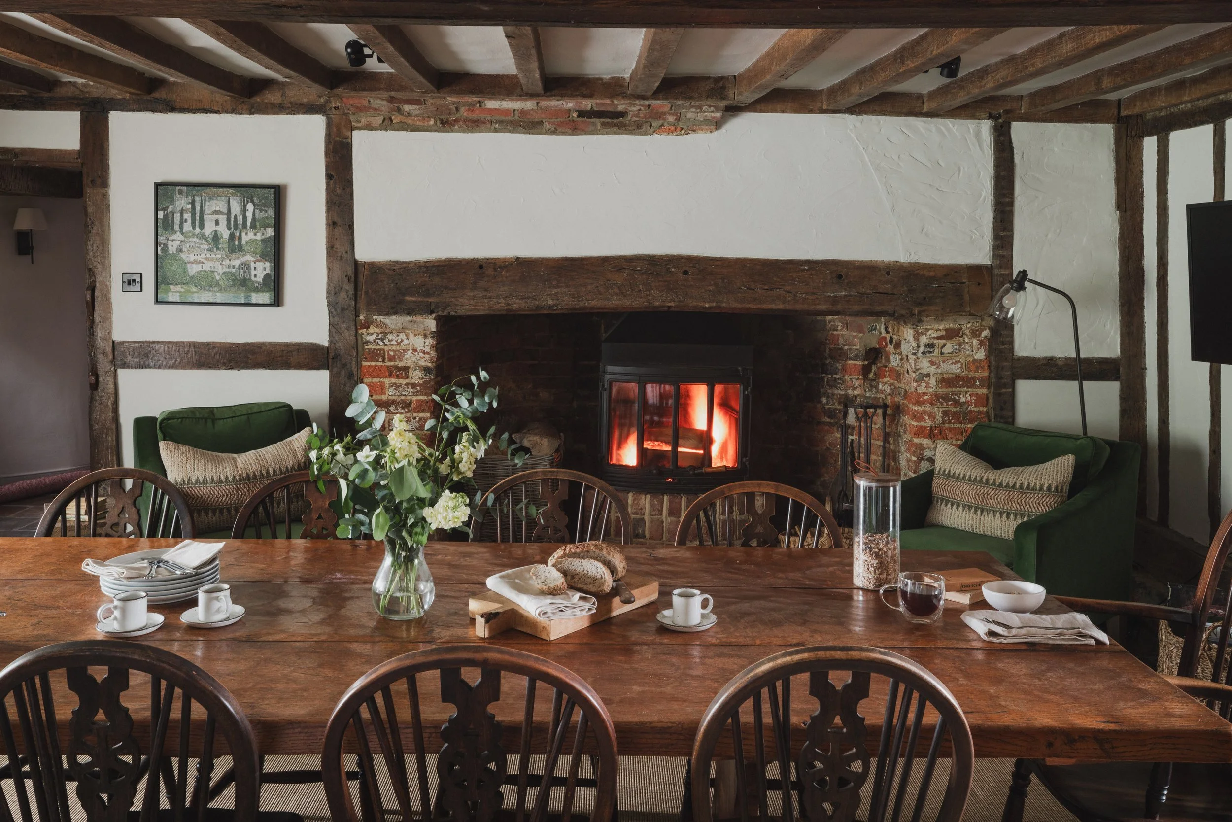 A cozy dining room with a rustic brick fireplace, wooden beams, and a wooden dining table set for breakfast with plates, cups, bread, and a flower vase.