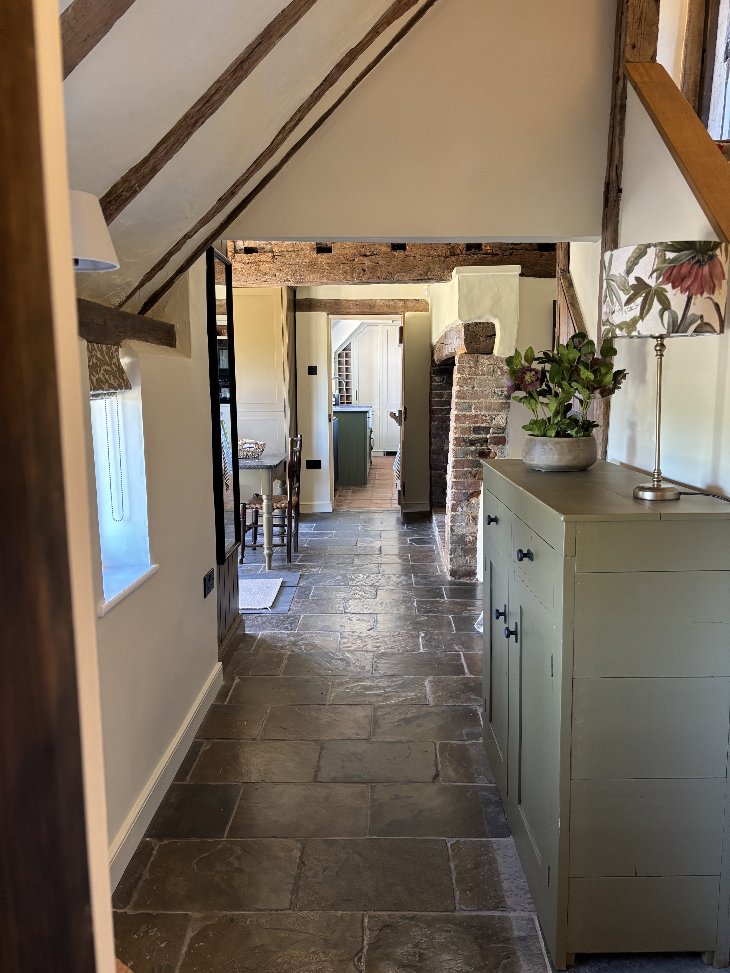 A view of an interior hallway with stone flooring, a cream-colored cabinet with a flower vase and lamp, exposed wooden beams, brick archway, wooden furniture, and a doorway leading to another room.
