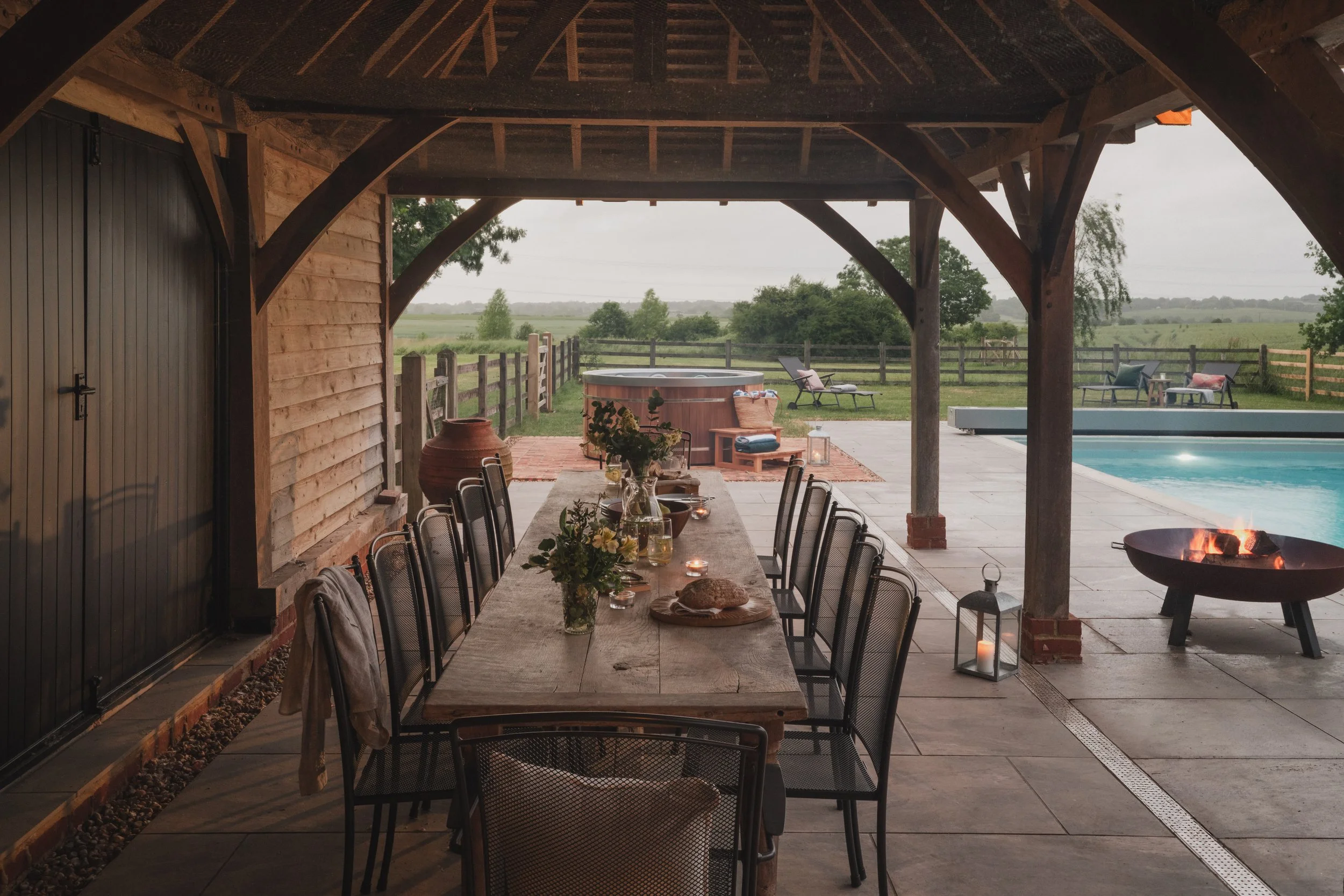 An outdoor patio and dining area under a wooden pergola, with a long dining table set with candles and decorative items, a fire pit, a hot tub, lounge chairs, and a pool, surrounded by a wooden fence and green landscape.