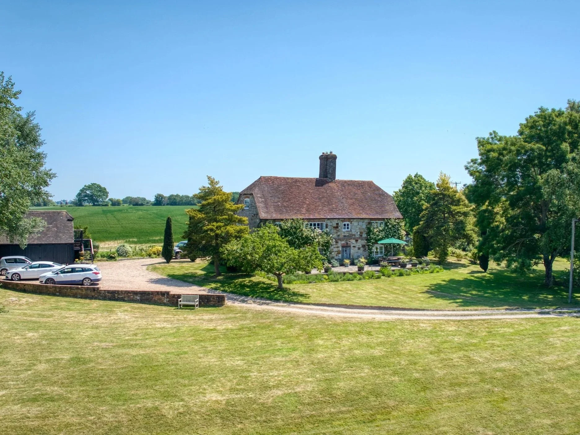 A stone house with a red-tiled roof and chimney surrounded by lush green trees and a well-maintained lawn, with parked cars and a gravel driveway in the foreground under a clear blue sky.