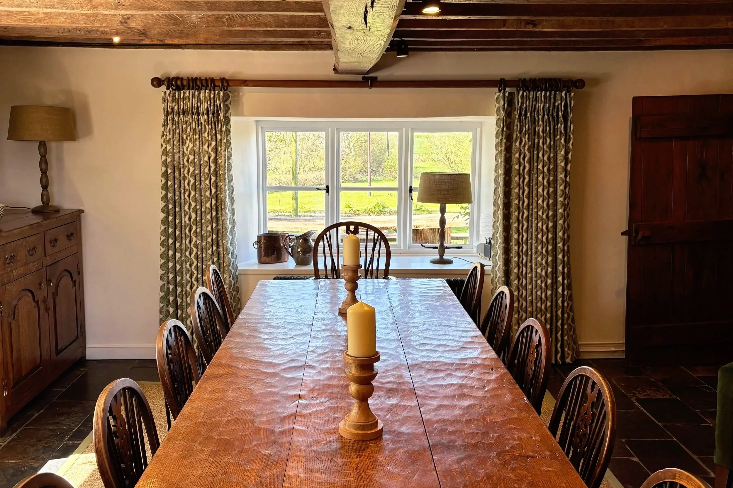 A wooden dining table with a tablecloth and three candles in the center, surrounded by several wooden chairs, in front of a window with patterned curtains, flanked by lamps on side tables, in a room with a wooden ceiling and furniture.